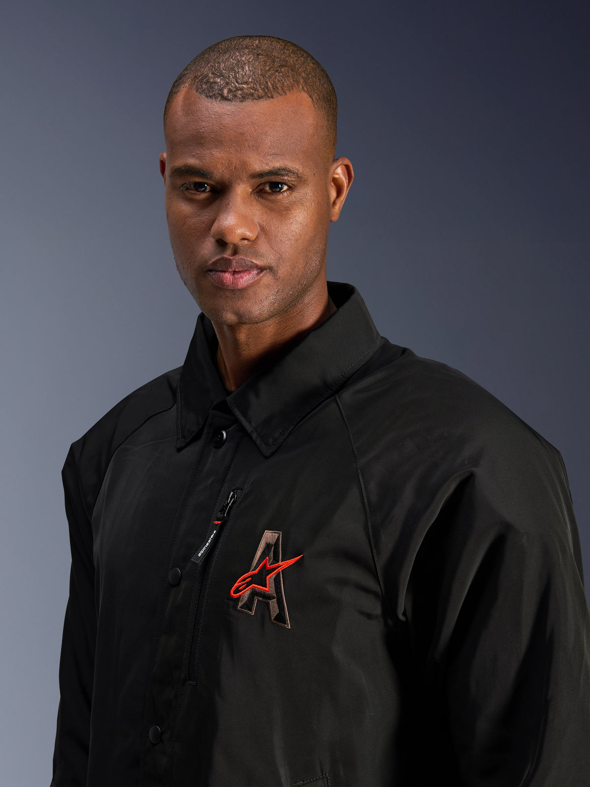 Alpinestars Revved WR Jacke, Schwarz, Nahaufnahme eines Mannes in der wasserabweisenden Satin-Motorradjacke, mit 3D-gesticktem „A“-Logo und rotem Alpinestars-Stern, Brusttasche mit Reißverschluss und Kragendesign für einen urbanen, zeitgenössischen Fahrstil.