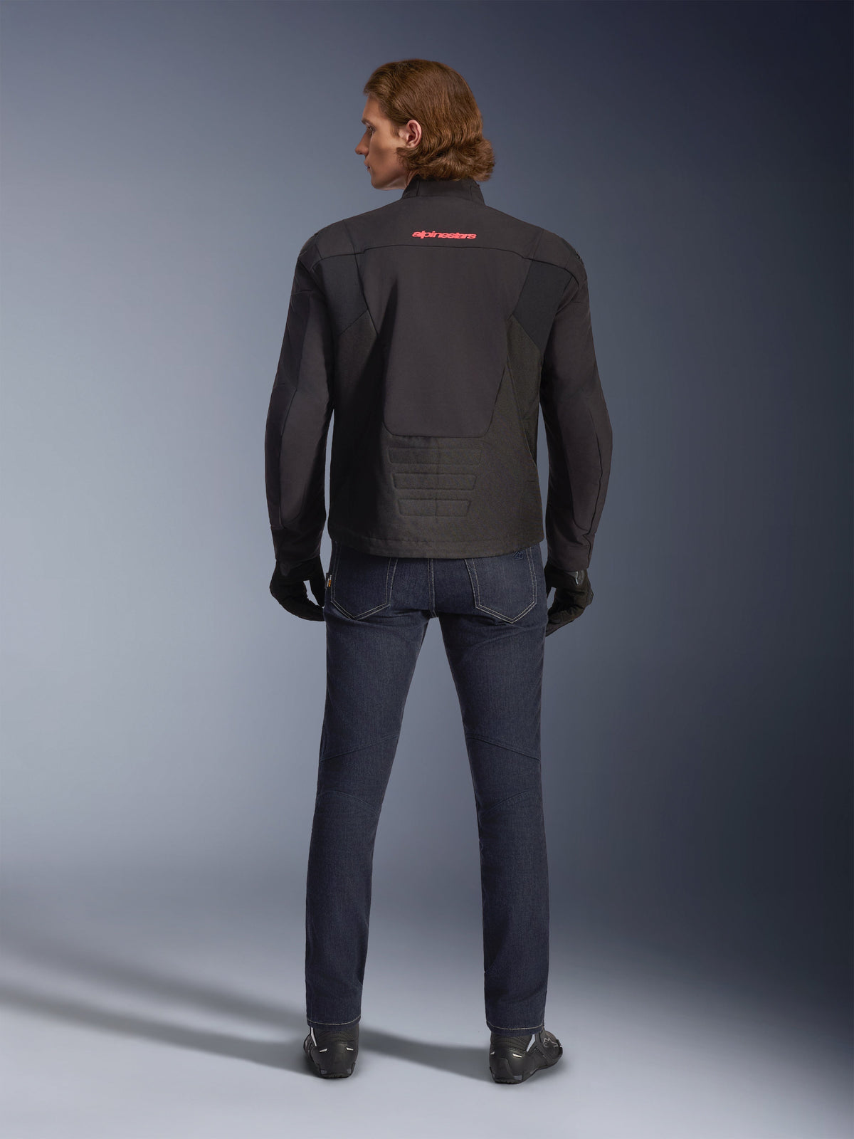 Alpinestars SMX Wasserdichte Jacke, Schwarz, Rückansicht getragen von einem Model, mit rotem Logo am oberen Rücken, verlängertem Rückensaum für optimale Abdeckung beim Fahren, kombiniert mit dunkelblauen Motorradjeans und schwarzen Handschuhen.