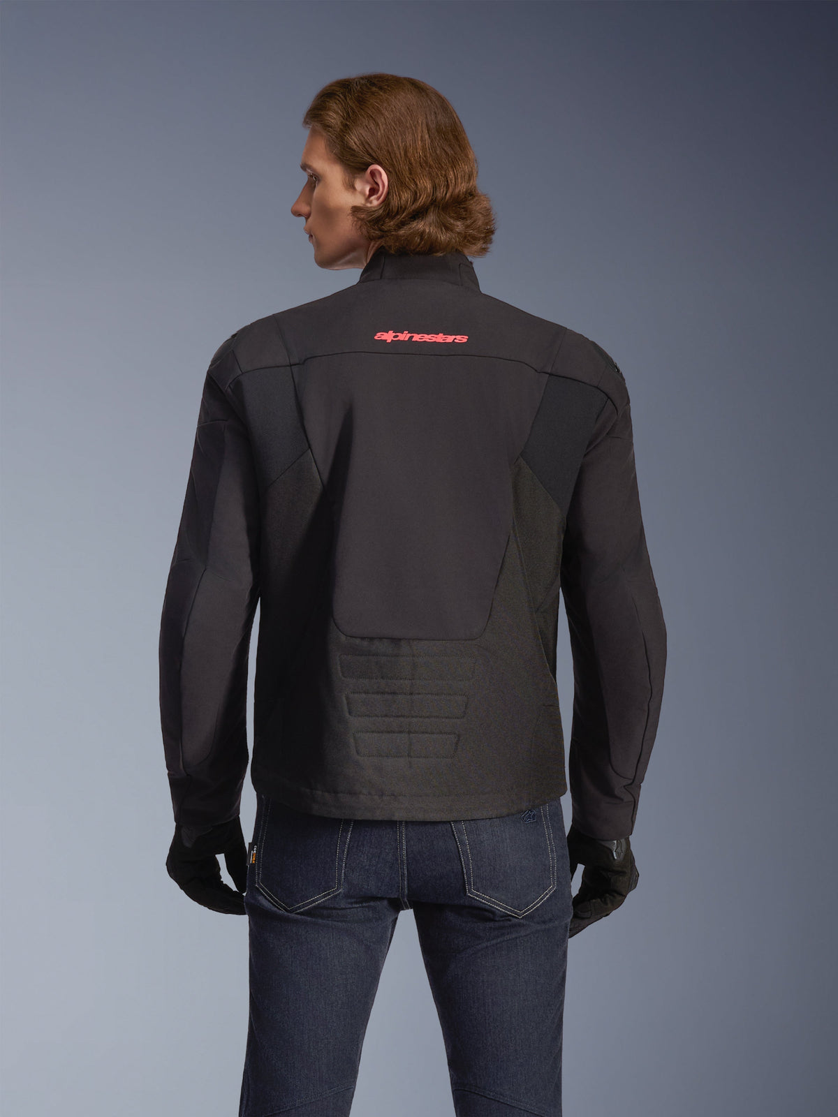 Alpinestars SMX Wasserdichte Jacke, Schwarz mit leuchtend rotem Logo, Rückansicht an einem Model, getragen mit dunkelblauen Jeans und schwarzen Handschuhen, Textil-Motorradjacke für Straßen- und Sportfahrten