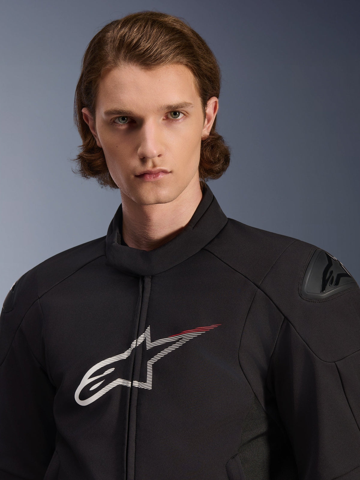 Modell trägt Alpinestars SMX Waterproof Jacket, Schwarz mit weißen und leuchtend roten Akzenten, großes weißes Stern-Logo mit rotem Streifen auf der Brust, DFS Lite Schulterprotektoren, schlanke Textilkonstruktion für Motorrad-Performance auf der Straße