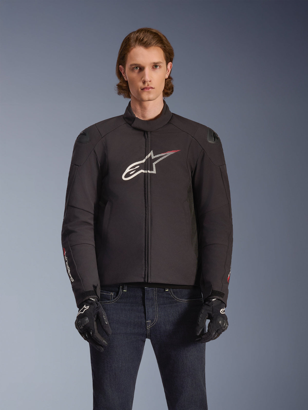 Alpinestars SMX Wasserdichte Jacke, schwarze Textil-Motorradjacke mit weißem und rotem Stern-Logo auf der Brust, getragen von einem Model mit schwarzen Handschuhen und blauen Jeans, mit externen Schulterschleifern und sportlicher Passform.