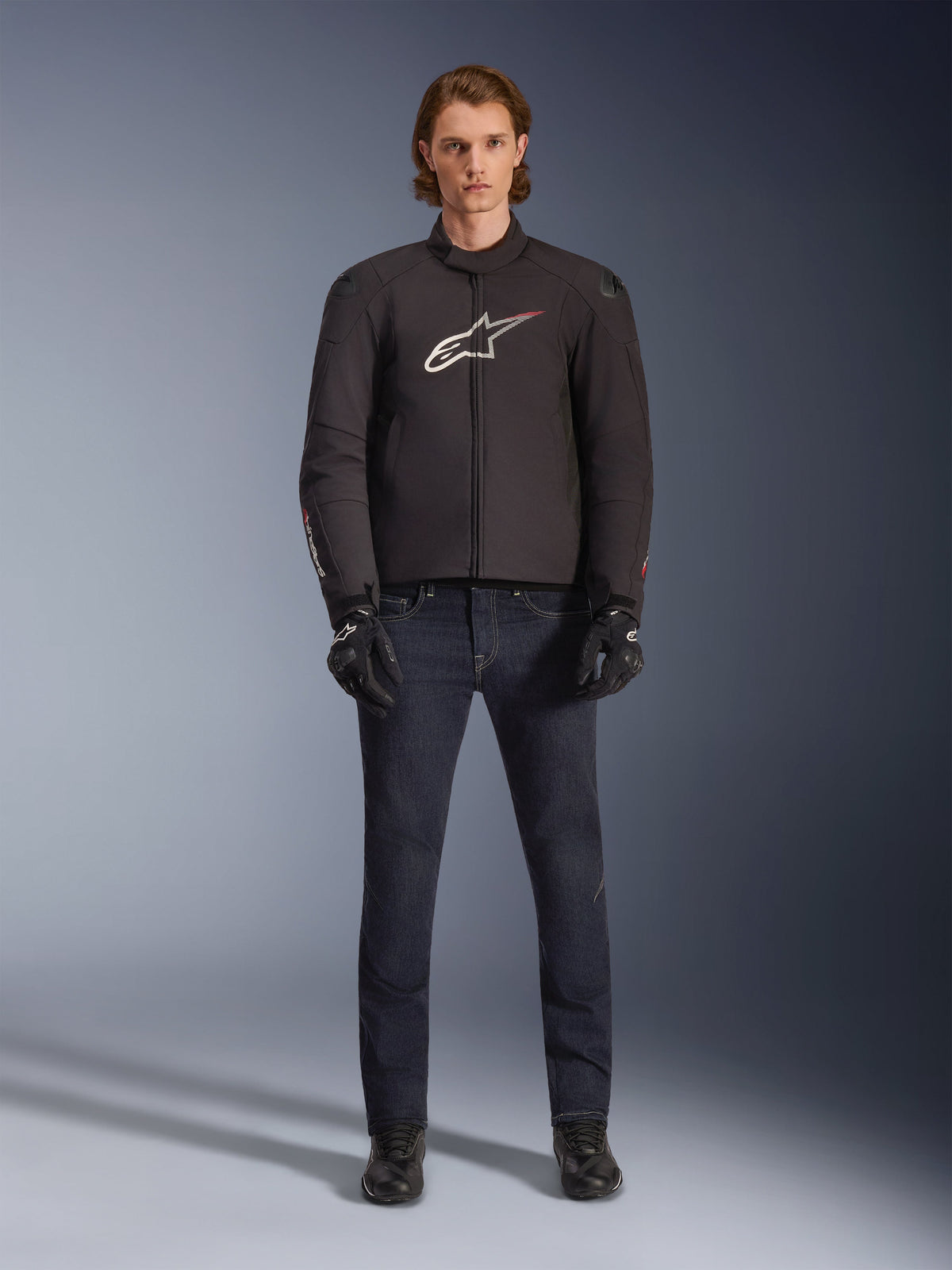 Model trägt die wasserdichte Alpinestars SMX Jacke in Schwarz mit weißem und rotem Logo, kombiniert mit einer dunklen Denim-Motorradhose, schwarzen Handschuhen und Motorradstiefeln, sportliches Allwetter-Motorrad-Outfit