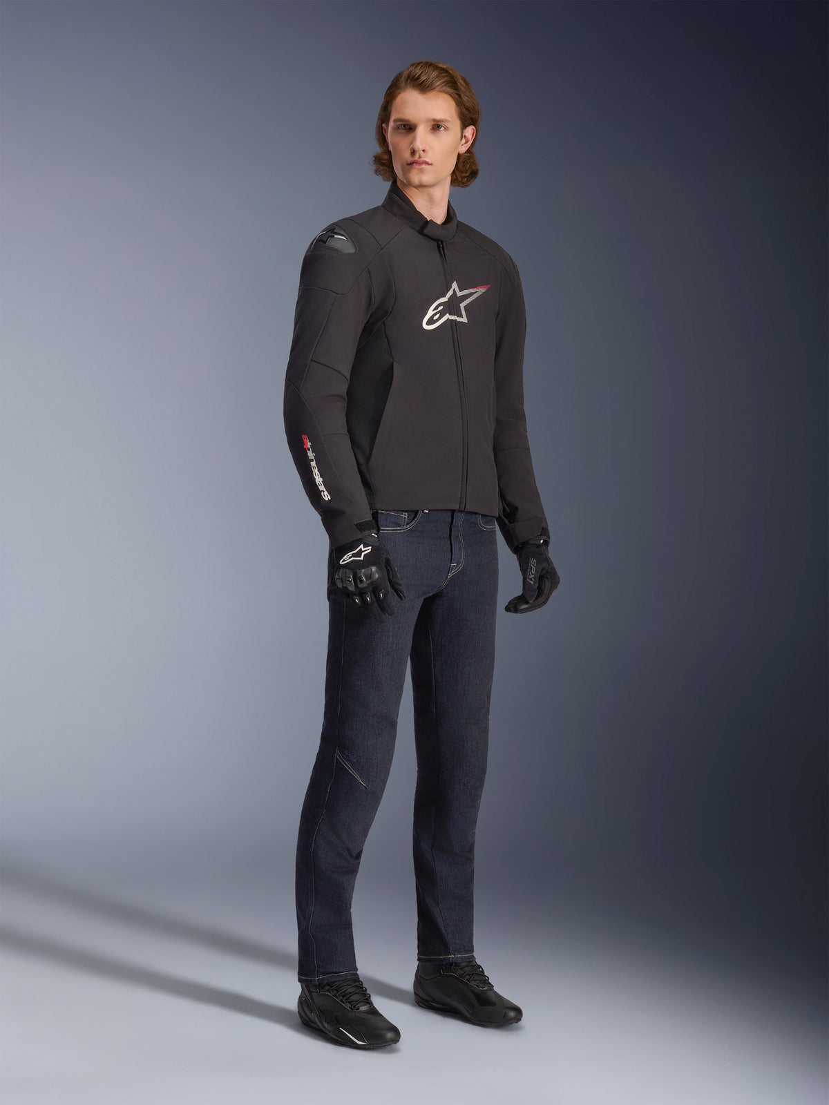 Männliches Model trägt Alpinestars SMX Waterproof Jacke, Schwarz mit weißen und roten Logo-Akzenten, kombiniert mit dunkelblauer Denim-Hose und schwarzen Motorradhandschuhen, schlanke Textilmotorradjacke mit sportlicher Passform.