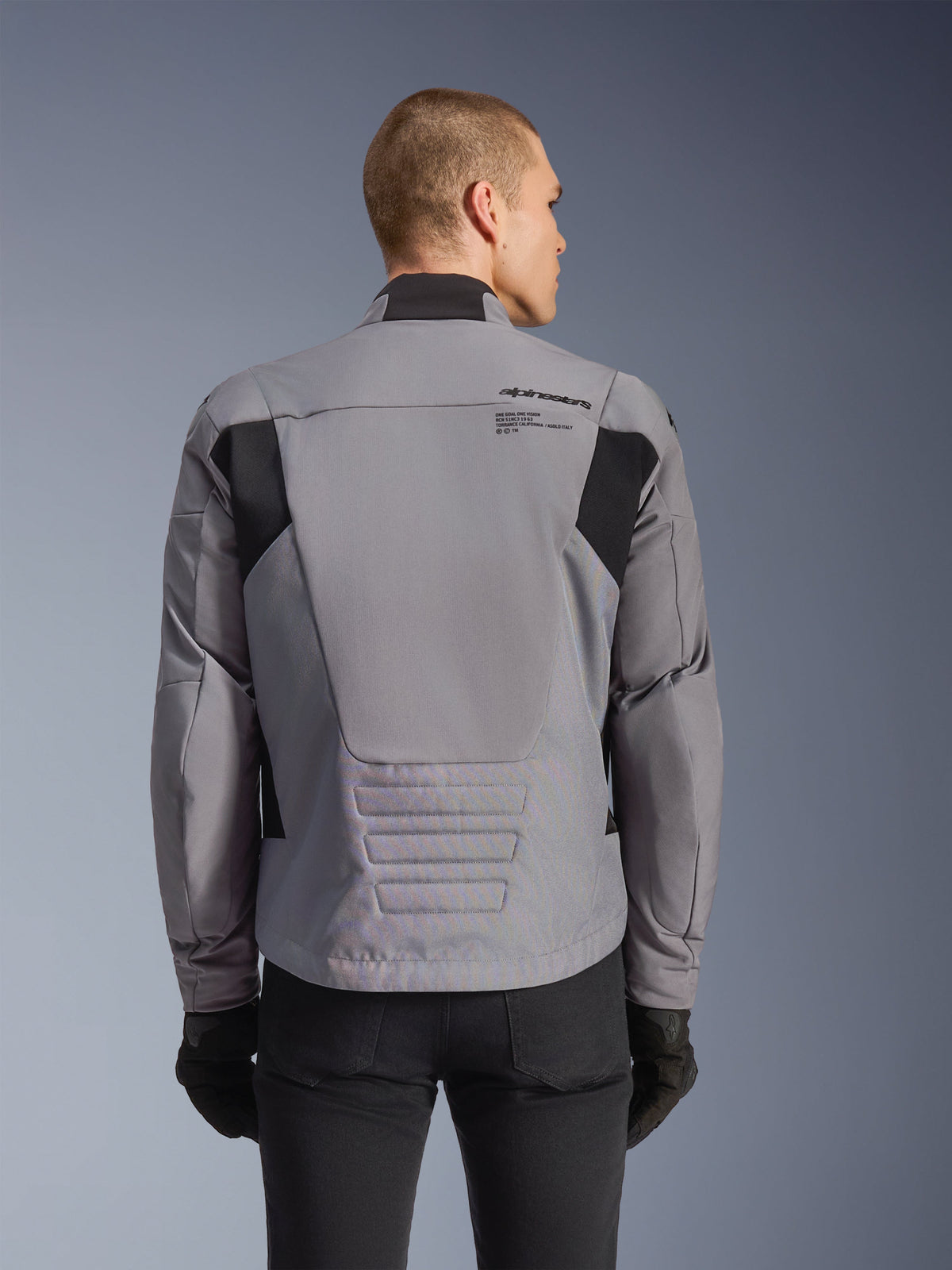 Alpinestars SMX Wasserdichte Jacke, Grau mit schwarzen Akzenten, Rückansicht mit Branding am oberen Rücken und verlängertem Profil, getragen von einem Model mit schwarzer Hose und Handschuhen