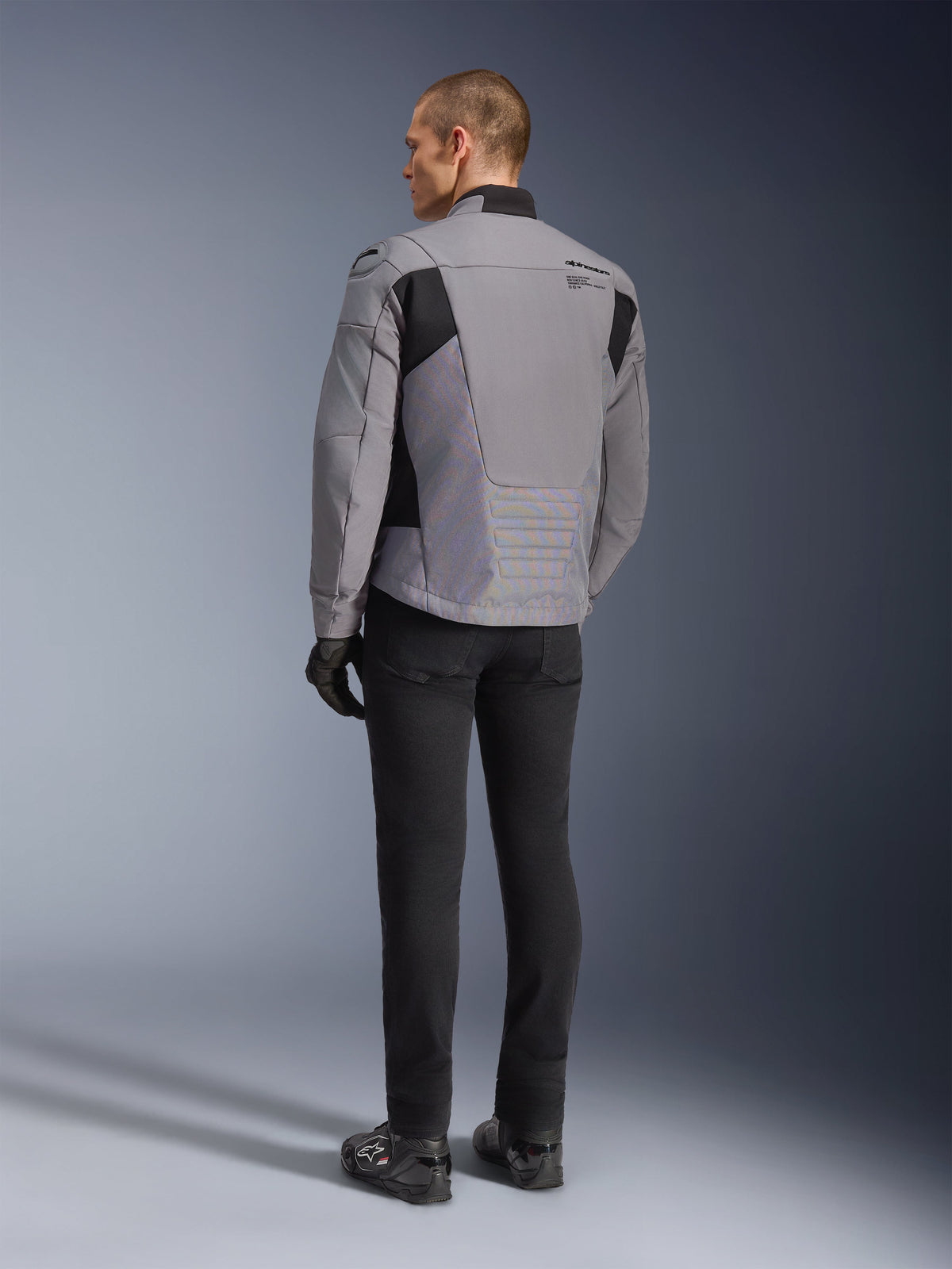 Alpinestars SMX Waterproof Jacke in Dunkelgrau und Schwarz, Rückansicht an einem Modell, getragen mit schwarzer Hose und Stiefeln, technische Textilkonstruktion mit schwarzen Einsätzen und Alpinestars-Branding auf der Schulter.