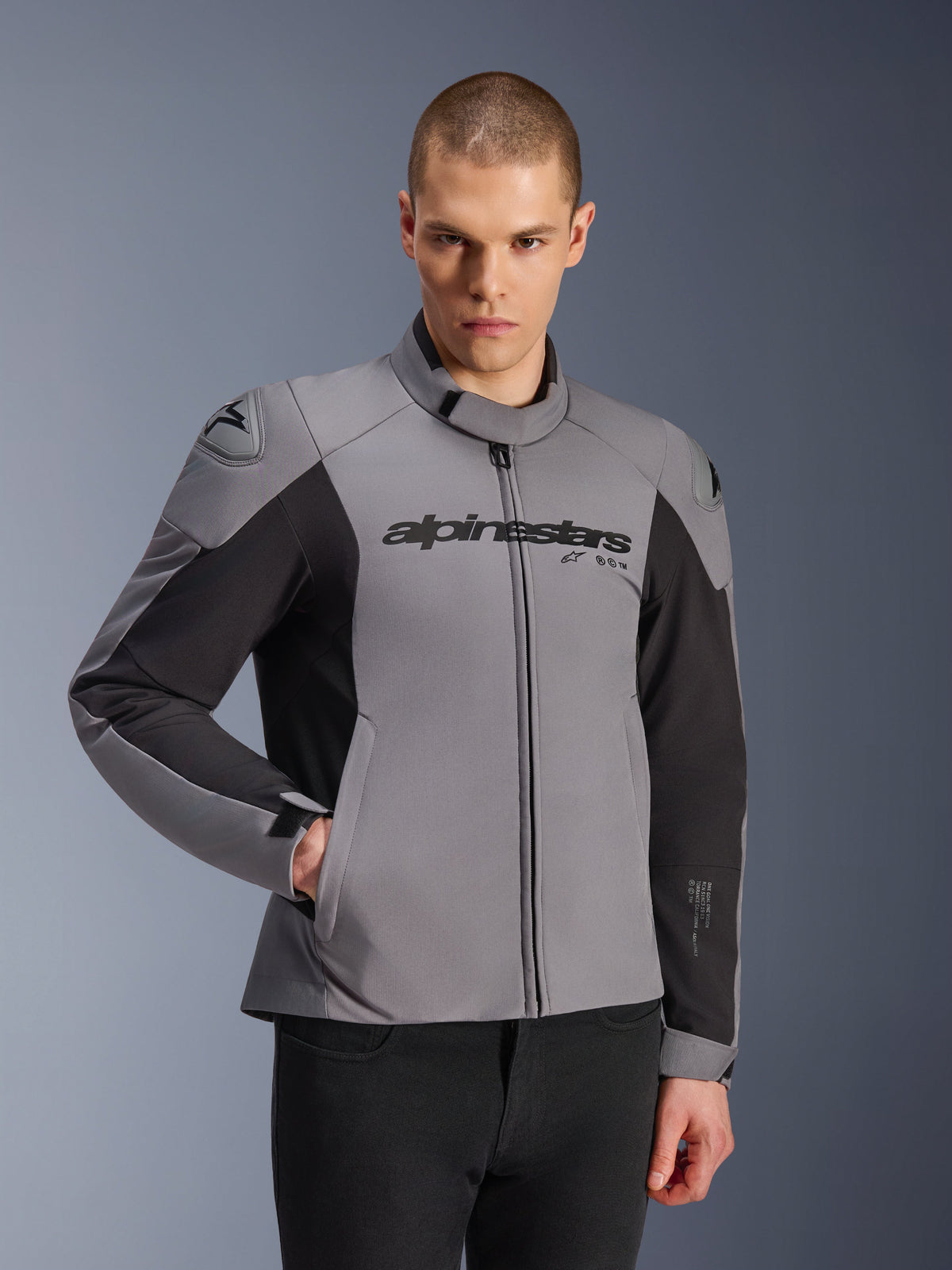 Alpinestars SMX Wasserdichte Jacke, Dunkelgrau und Schwarz, getragen von einem männlichen Model, mit „Alpinestars“-Branding auf der Brust, DFS Lite Schulterschutz, schlankes sportliches Design aus Textil- und Softshell-Materialien für Straßenfahrten.