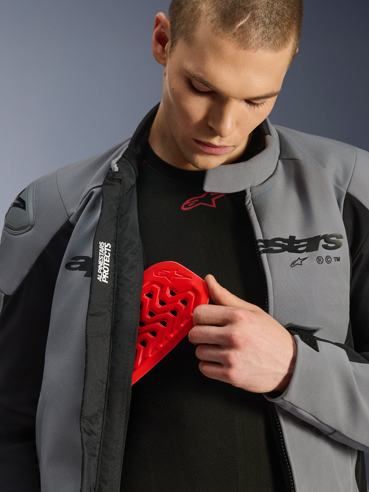 Männliches Model in der Alpinestars SMX Waterproof Jacke in Dark Gray Black, das einen roten Nucleon-Brustprotektor in eine Innentasche einsetzt, um die Protektorenkompatibilität und Schutzeigenschaften der Jacke zu zeigen.