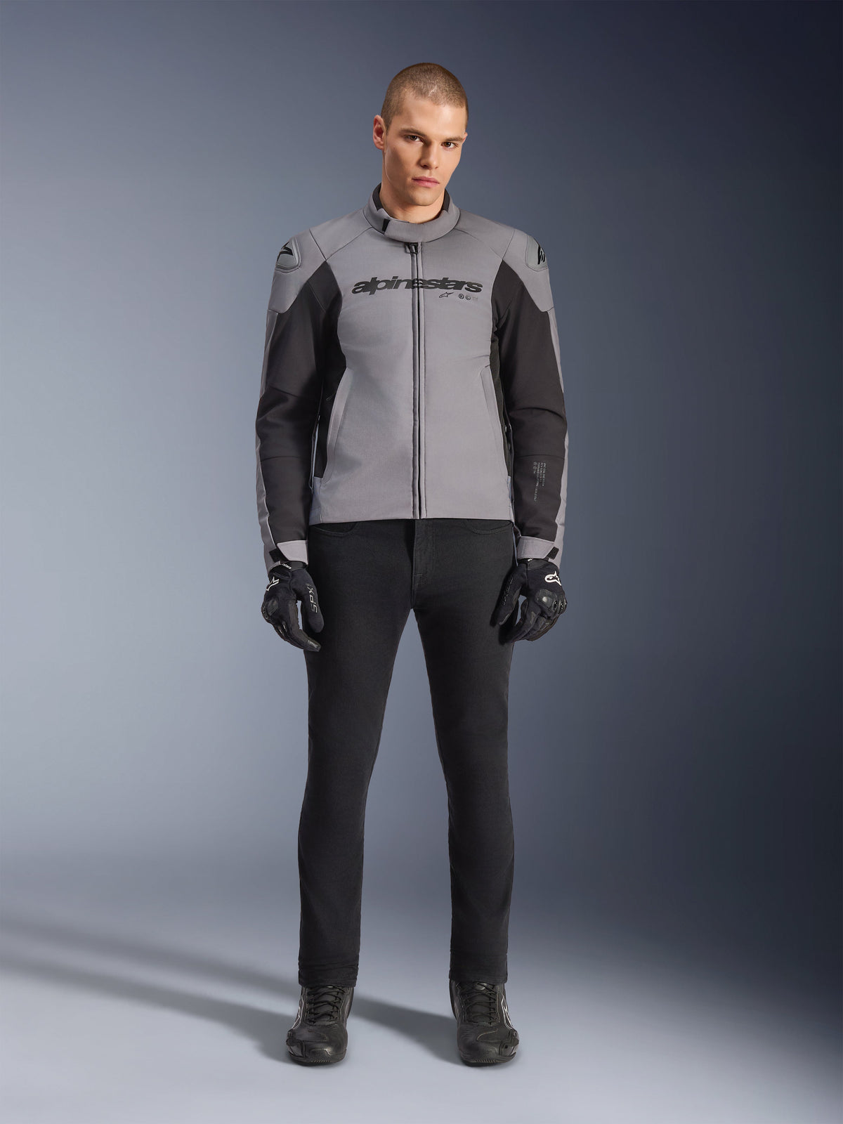 Alpinestars SMX Waterproof Jacke, Dunkelgrau und Schwarz, Frontansicht an einem männlichen Model, sportliche Textil-Motorradjacke mit schwarzen Seiteneinsätzen und Alpinestars-Branding auf der Brust, kombiniert mit schwarzer Motorradhose und Handschuhen.