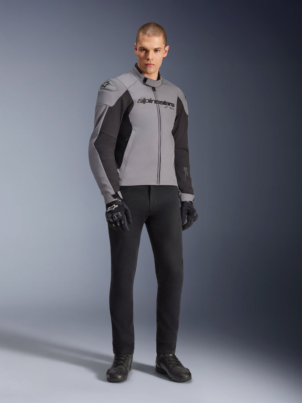 Alpinestars SMX Wasserdichte Jacke, Motorradjacke, Dunkelgrau und Schwarz, getragen von einem Model mit schwarzen Handschuhen und schwarzer Hose, mit markantem Branding auf der Brust und einem technischen, sportbetonten Design für Straßenfahrten bei jedem Wetter.