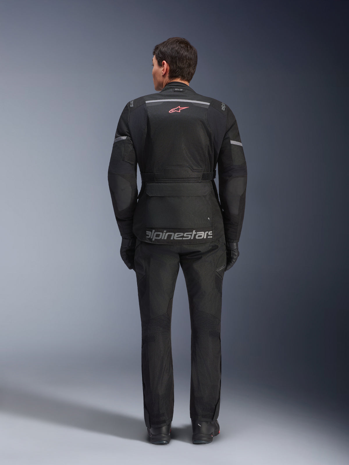 Alpinestars Andes Air Drystar® Jacke und Hose, schwarzes Motorrad-Tourenoutfit, Rückansicht des Fahrers, mit Mesh-Einsätzen, rotem Stern-Logo und reflektierendem Alpinestars-Branding auf der Jacke, getragen mit schwarzen Handschuhen und Stiefeln.