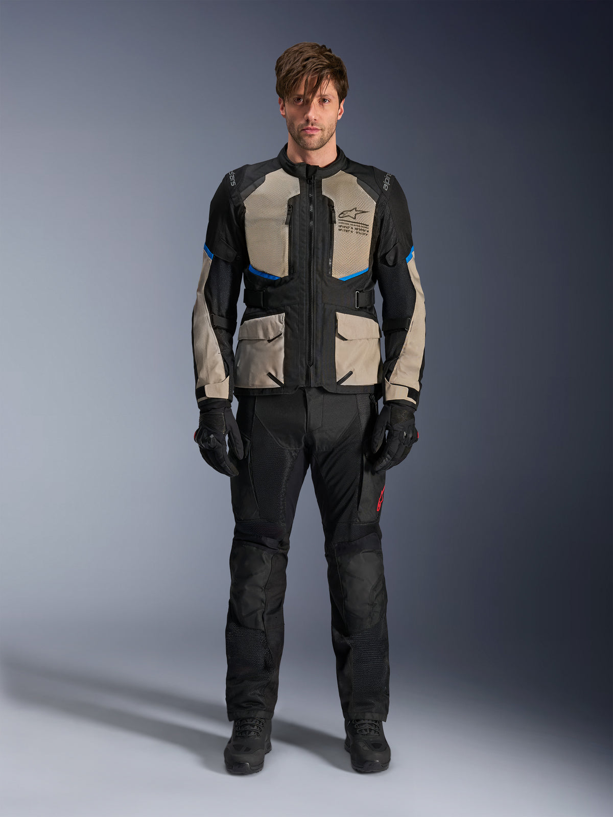 Modell trägt Alpinestars Andes Air Drystar® Jacke, Motorrad-Tourenjacke, Schwarz, Laurel Gray und Bright Blue, kombiniert mit passender schwarzer Fahrhose und Handschuhen, mit großflächigen Mesh-Belüftungseinsätzen an Brust und Ärmeln für Adventure-Touren bei warmem Wetter.