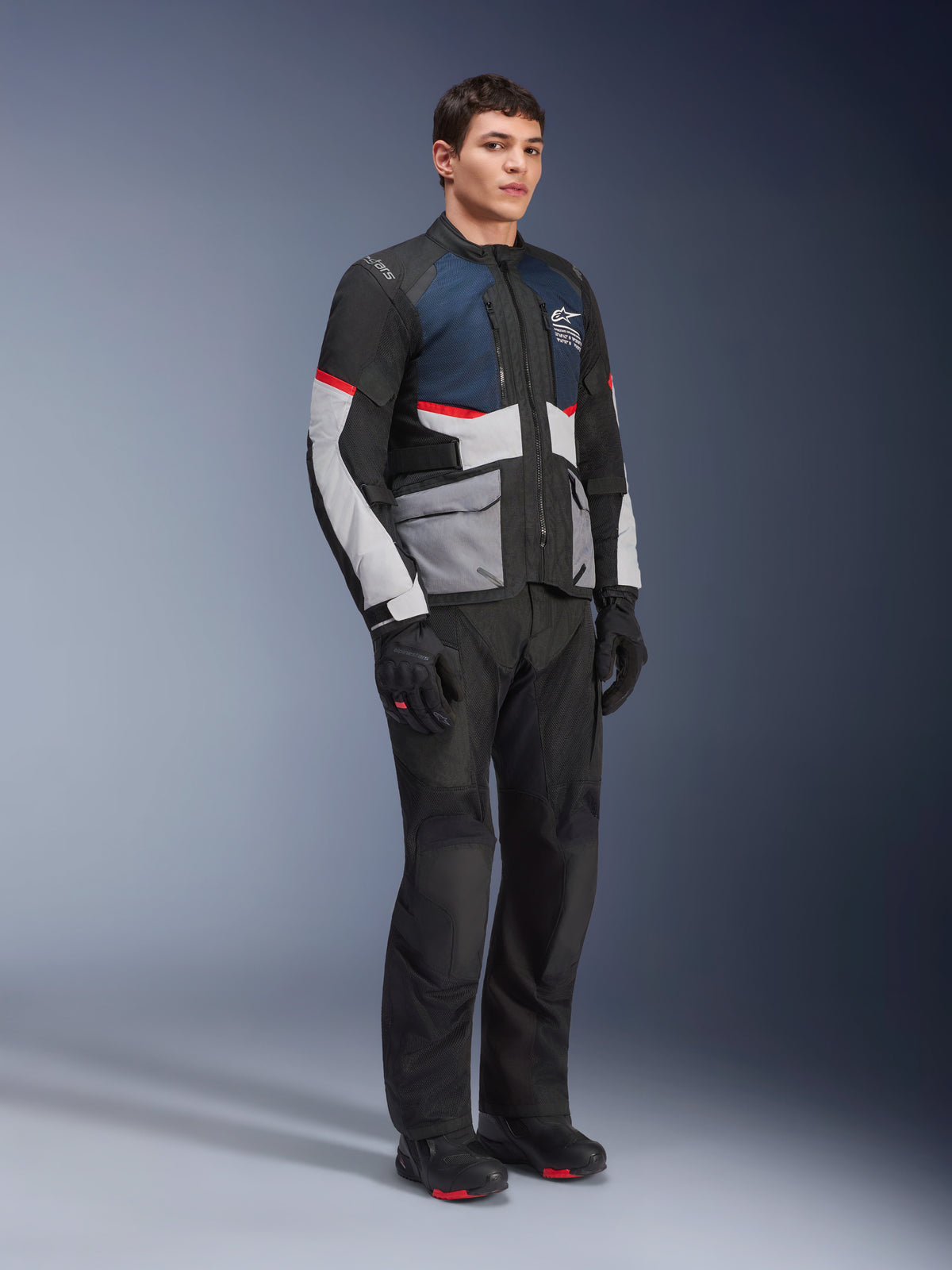 Männliches Model trägt die Alpinestars Andes Air Drystar® Jacke in Deep Blue Black Ice Gray, kombiniert mit schwarzer Motorradhose, Handschuhen und Stiefeln, mit großen Mesh-Einsätzen zur Belüftung, hellgrauen Taschen und roten Akzenten, entworfen für Adventure-Touring.