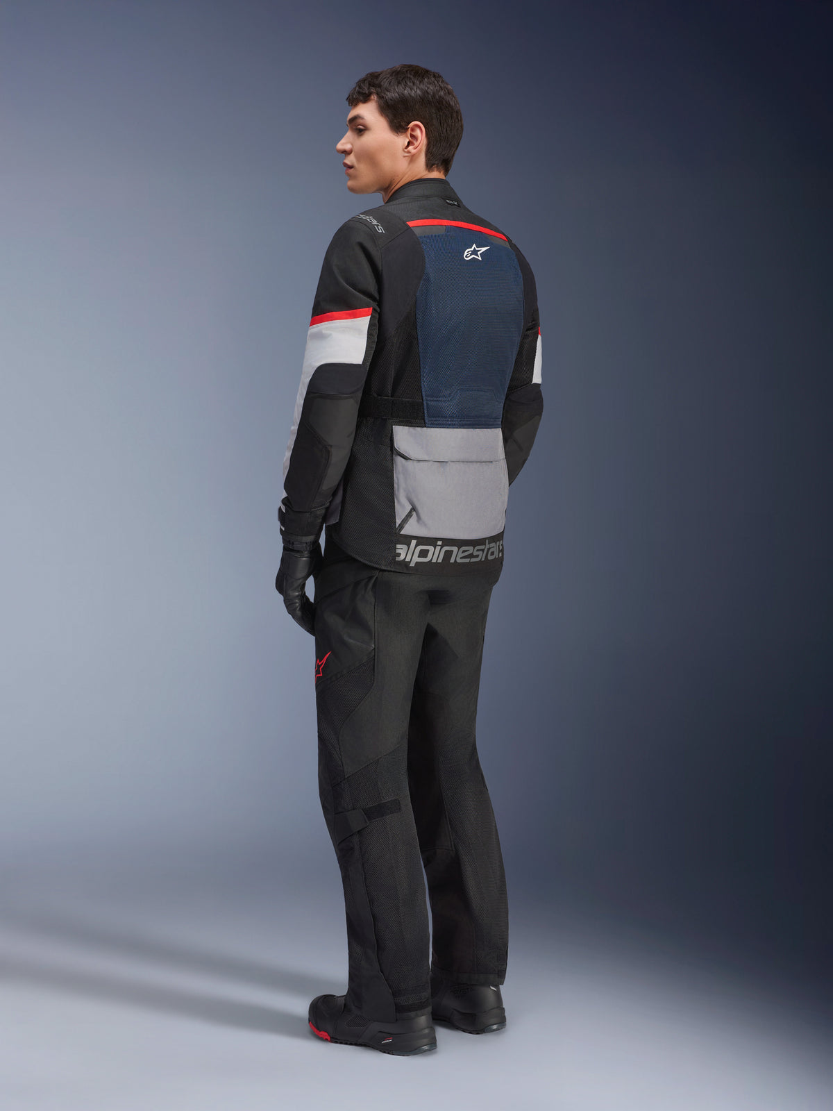 Männliches Model in der Rückansicht von der Seite, das die Alpinestars Andes Air Drystar® Jacke in Deep Blue Black Ice Gray trägt, kombiniert mit schwarzer Motorradhose und Handschuhen, mit blauem Mesh-Rückeneinsatz, grauer Utility-Tasche am unteren Rücken und roten Akzenten an den Ärmeln, professionelle Adventure-Touring-Ausrüstung.