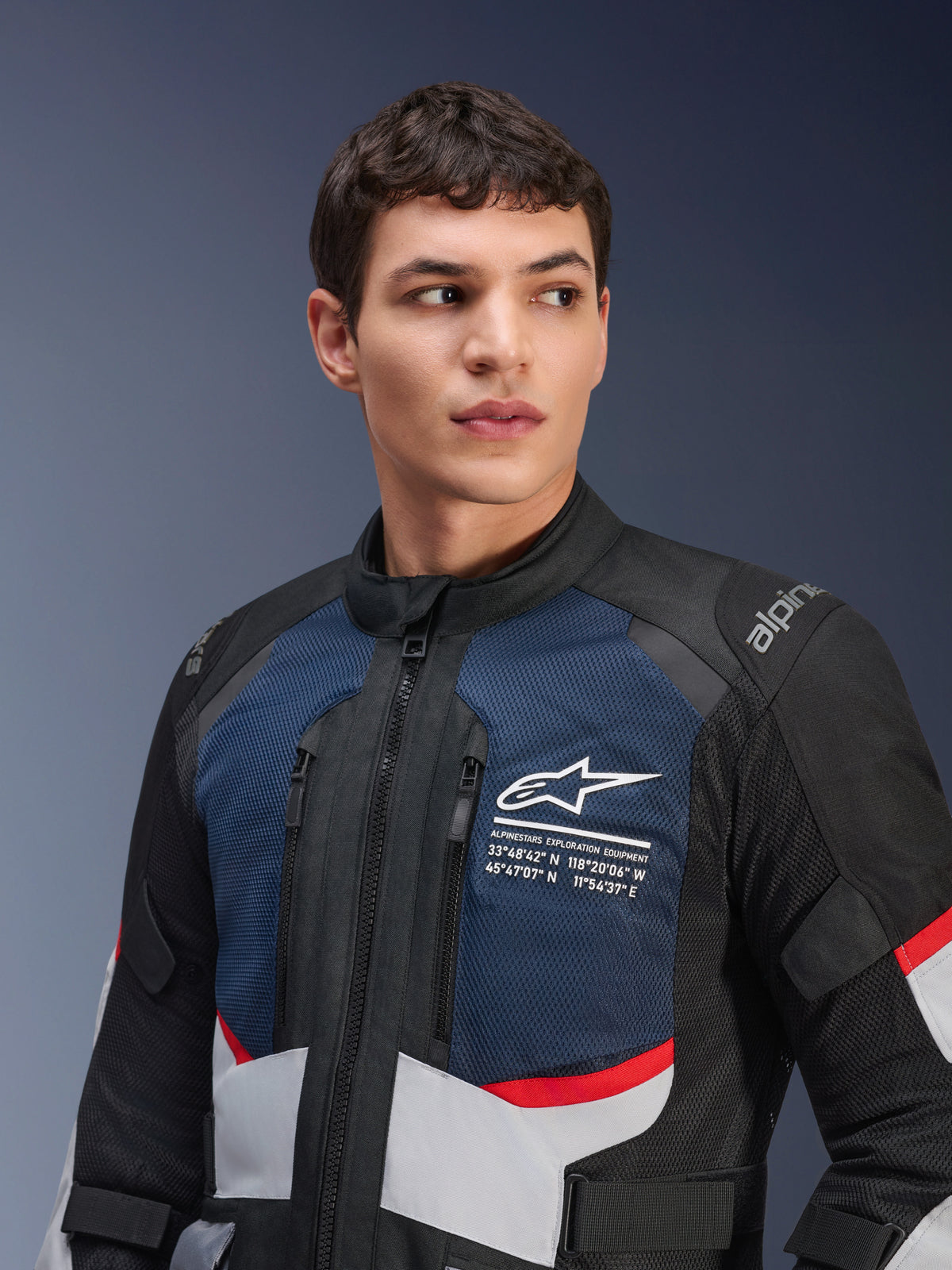 Alpinestars Andes Air Drystar® Jacke, Motorradjacke, Tiefblau, Schwarz und Eisgrau mit roten Akzenten, getragen von einem männlichen Model, mit belüfteten Mesh-Brustpaneelen, weißem Stern-Logo mit Explorations-Koordinaten und straßentauglichem Adventure-Design