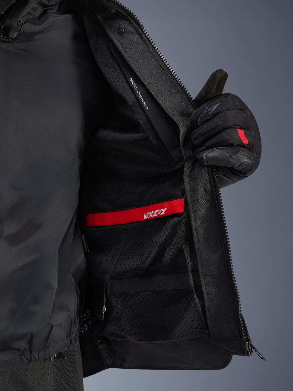 Innen-Detailansicht der Alpinestars Andes Air Drystar® Jacke, zeigt das atmungsaktive schwarze Mesh-Futter, das rote wasserdichte Fach und die Brustprotektortasche, die von einem schwarzen Motorradhandschuh offen gehalten wird.