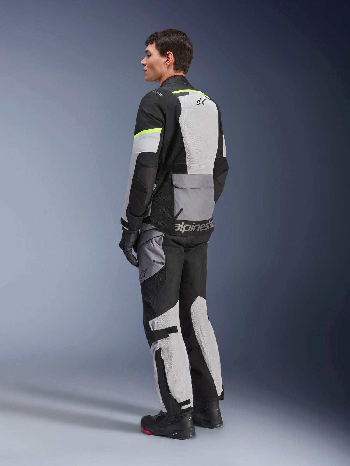 Alpinestars Andes Air Drystar® Jacke, Touren-Motorradjacke, Eisgrau und Dunkelgrau mit schwarzen Einsätzen und neongelben Akzenten, getragen mit passender Hose und Handschuhen, Dreiviertel-Rückansicht mit Mesh-Belüftung und Alpinestars-Branding.