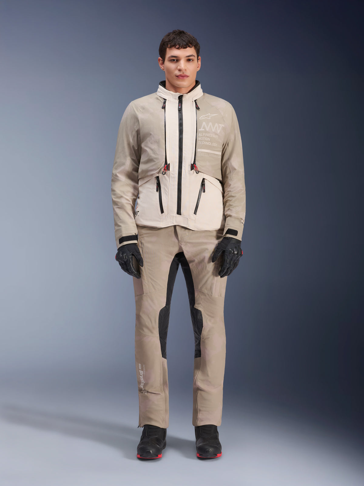 Alpinestars AMT-10LAB Drystar® XF Jacke in White Sand Aluminum Camo, getragen von einem Model mit passender Adventure-Fahrhose, schwarzen Handschuhen und schwarzen Stiefeln, technische modulare Motorradausrüstung für Multisaison-Touren.