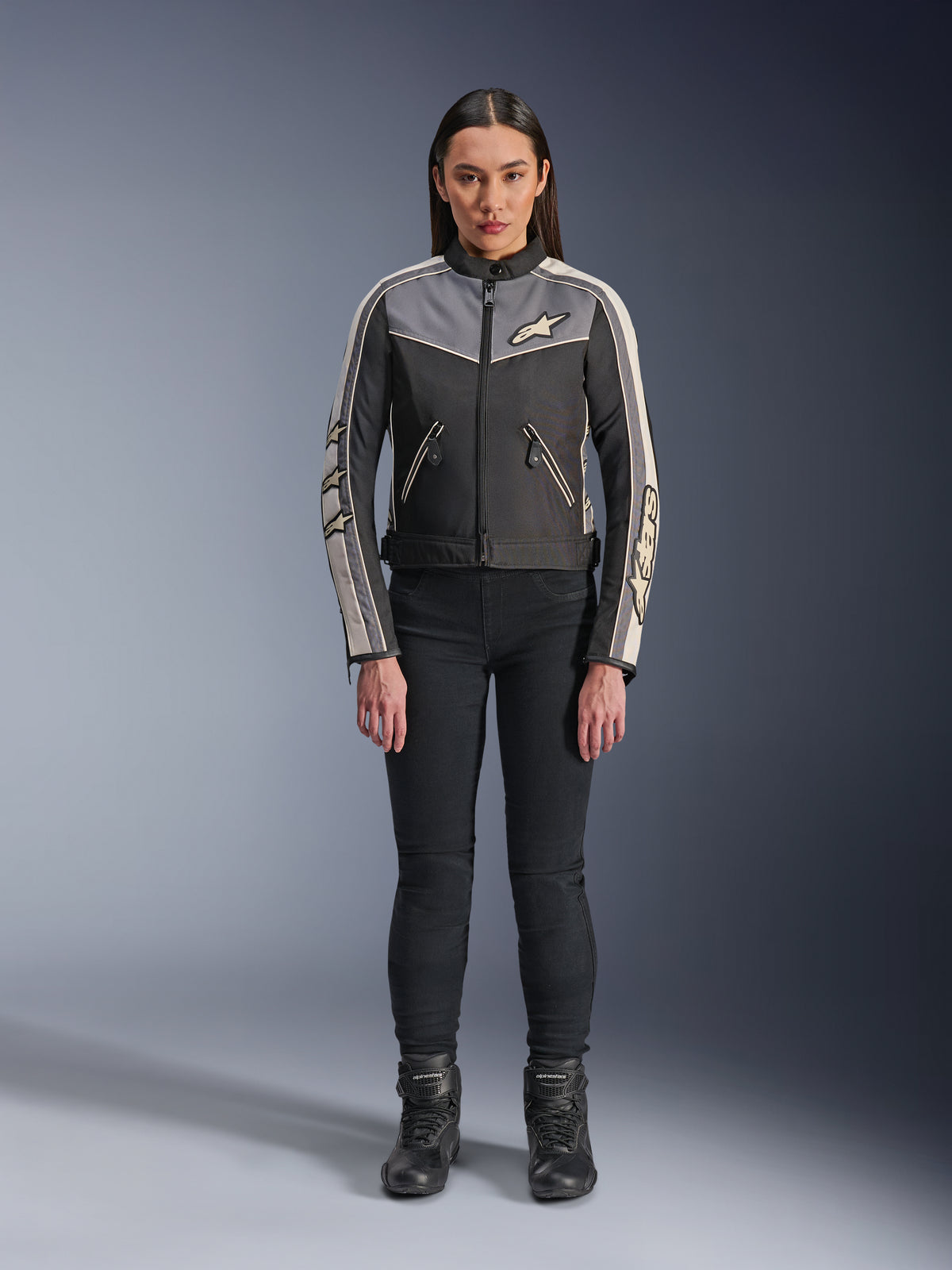 Frau in Alpinestars Stella T-Dyno WR Jacke, Schwarz, Dunkelgrau und Ecru, Vorderansicht, Textil-Motorradjacke mit vintage-inspiriertem Design, Branding an Brust und Ärmeln, getragen mit schwarzer Hose und Stiefeln