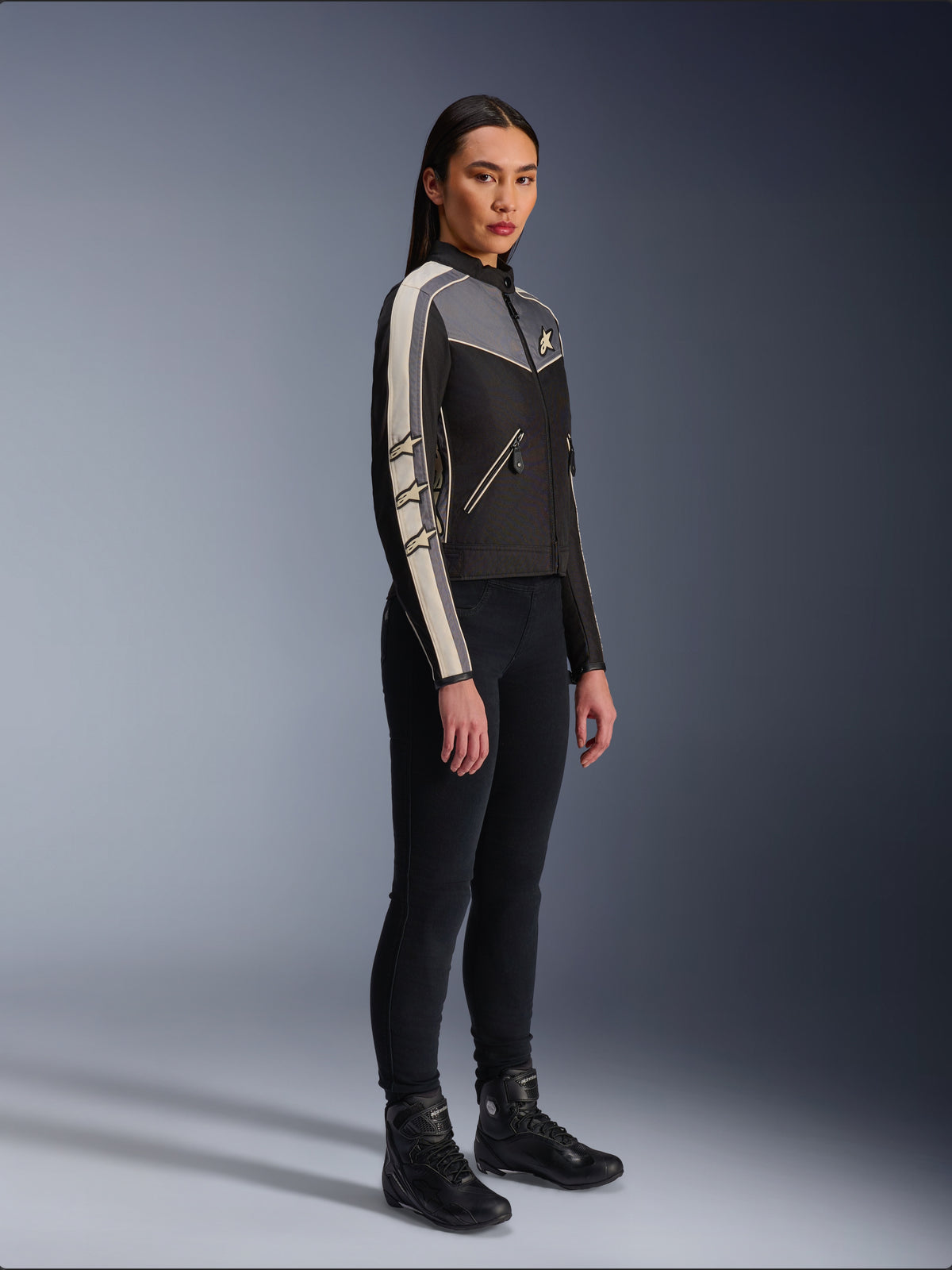 Alpinestars Stella T-Dyno WR Jacke, Textil-Motorradjacke für Damen, Schwarz, Dunkelgrau und Ecru, Vintage-inspiriertes Design mit cremefarbenen Ärmelstreifen und Stern-Logos, getragen mit schwarzer Hose und Motorradstiefeln, Heritage-inspirierte Urban-Riding-Ausrüstung