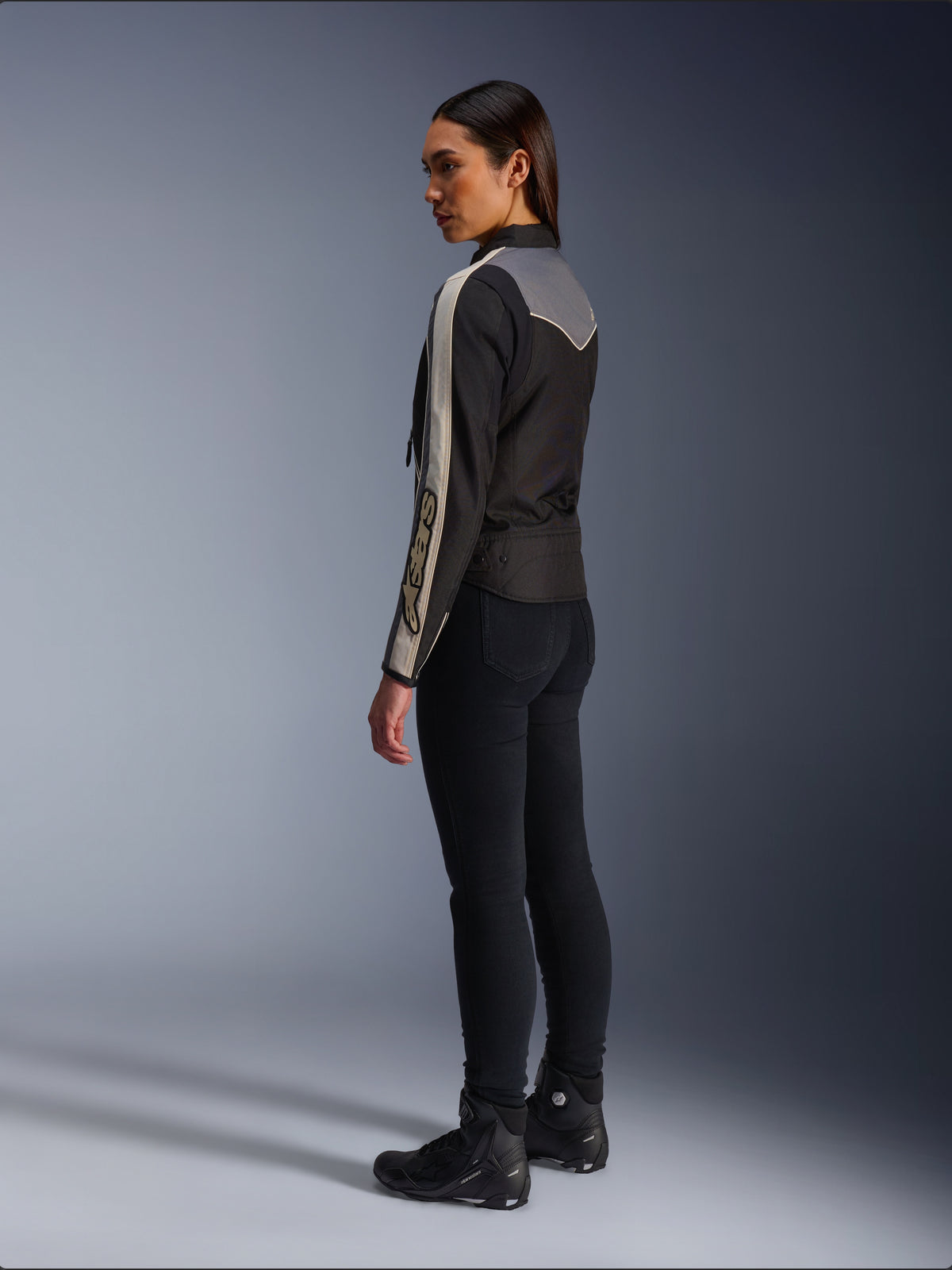 Weibliches Model in der Seiten- und Rückansicht, trägt die Alpinestars Stella T-Dyno WR Jacke in Schwarz Dunkelgrau Ecru, mit vintage-inspiriertem Design, ecrufarbenen Ärmelstreifen und grauer Rückenpartie, kombiniert mit schwarzer Hose und schwarzen Motorradschuhen.