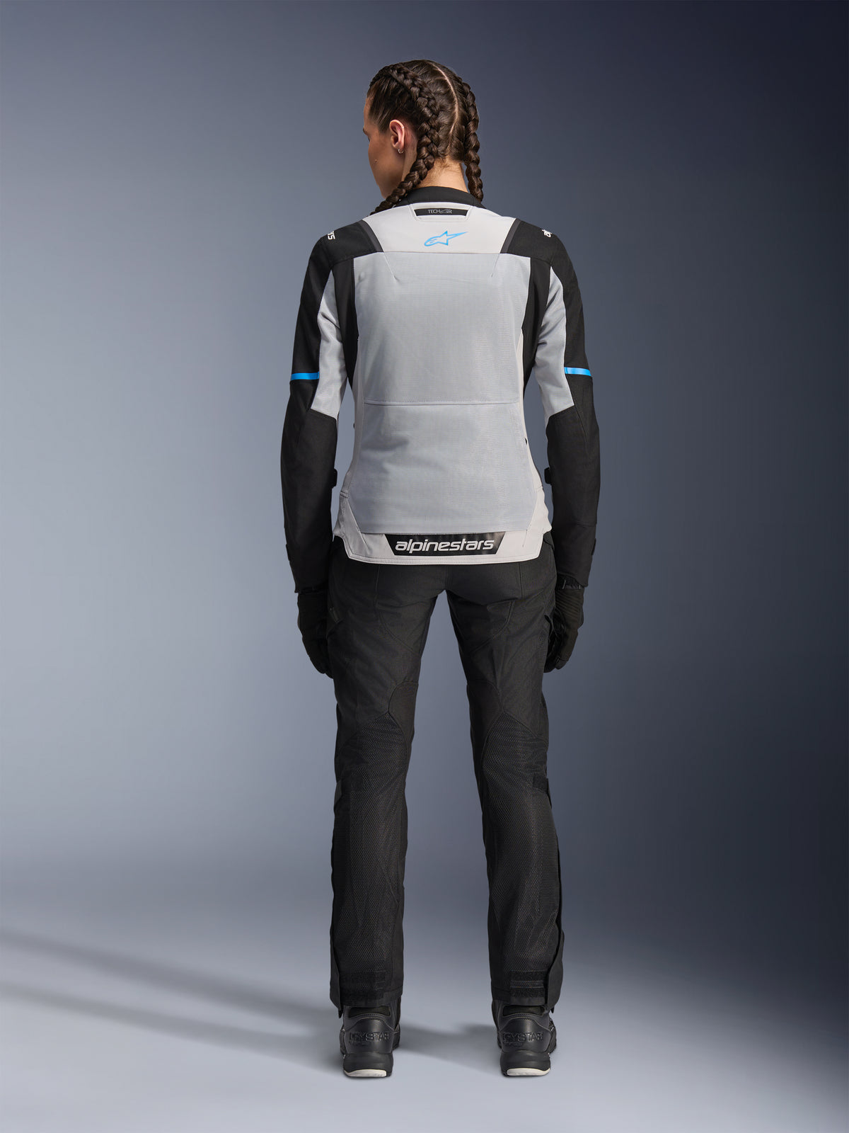 Alpinestars Stella ST-2 Air Jacke, Ice Gray Black, Rückansicht an weiblichem Model, mit großem hellgrauem Mesh-Rückenpanel, schwarzen Ärmeln mit blauen Akzentstreifen, Alpinestars-Branding am unteren Rücken, kombiniert mit schwarzer technischer Motorradhose und Stiefeln, Sommer-Motorrad-Tourenausrüstung