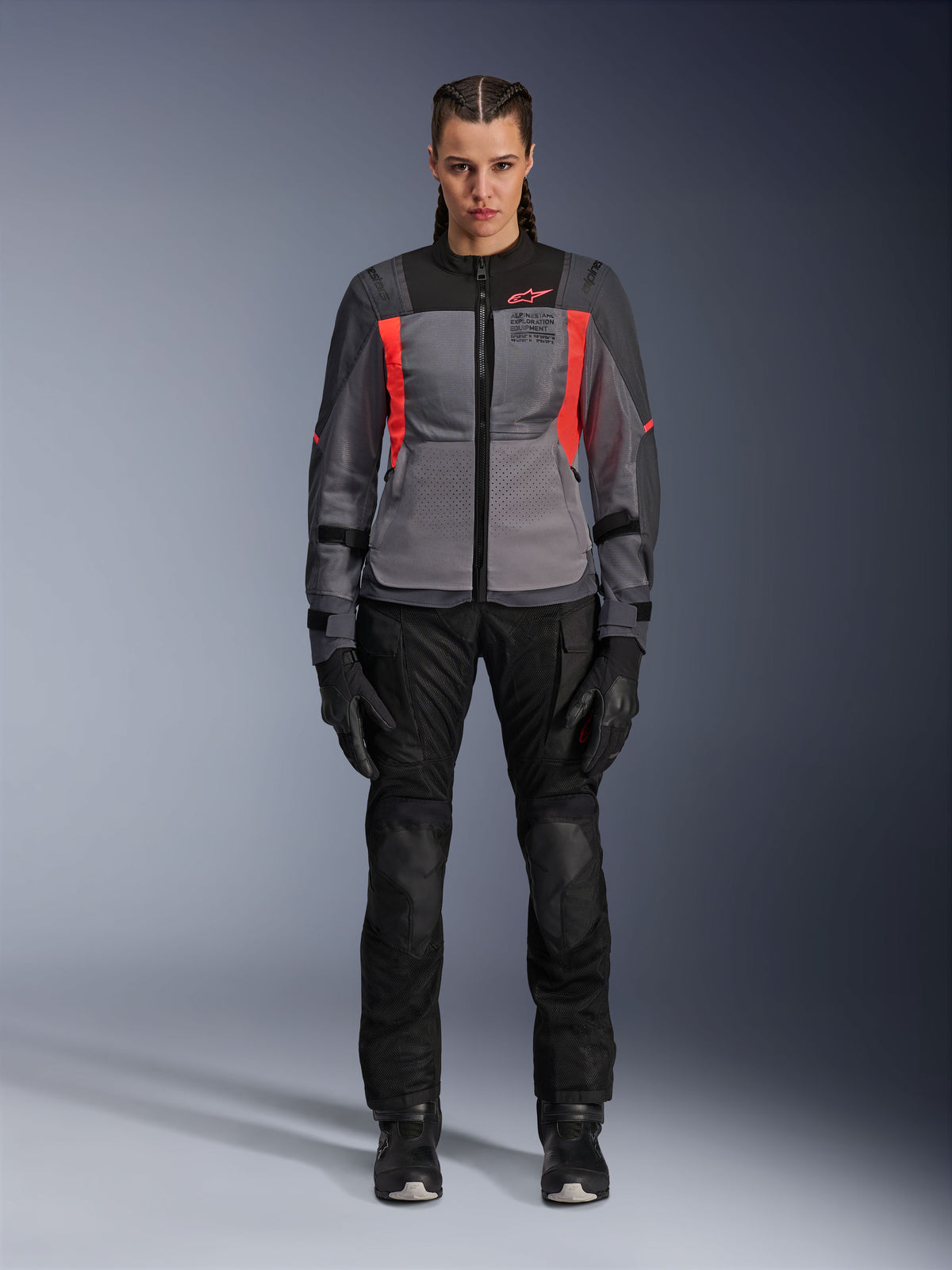 Frau trägt Alpinestars Stella ST-2 Air Jacke in Deep Gray, Dark Gray und Fluo Coral, kombiniert mit schwarzer Motorradhose und Handschuhen, Frontansicht, moderne Mesh-Adventure-Tourenausrüstung für Damen