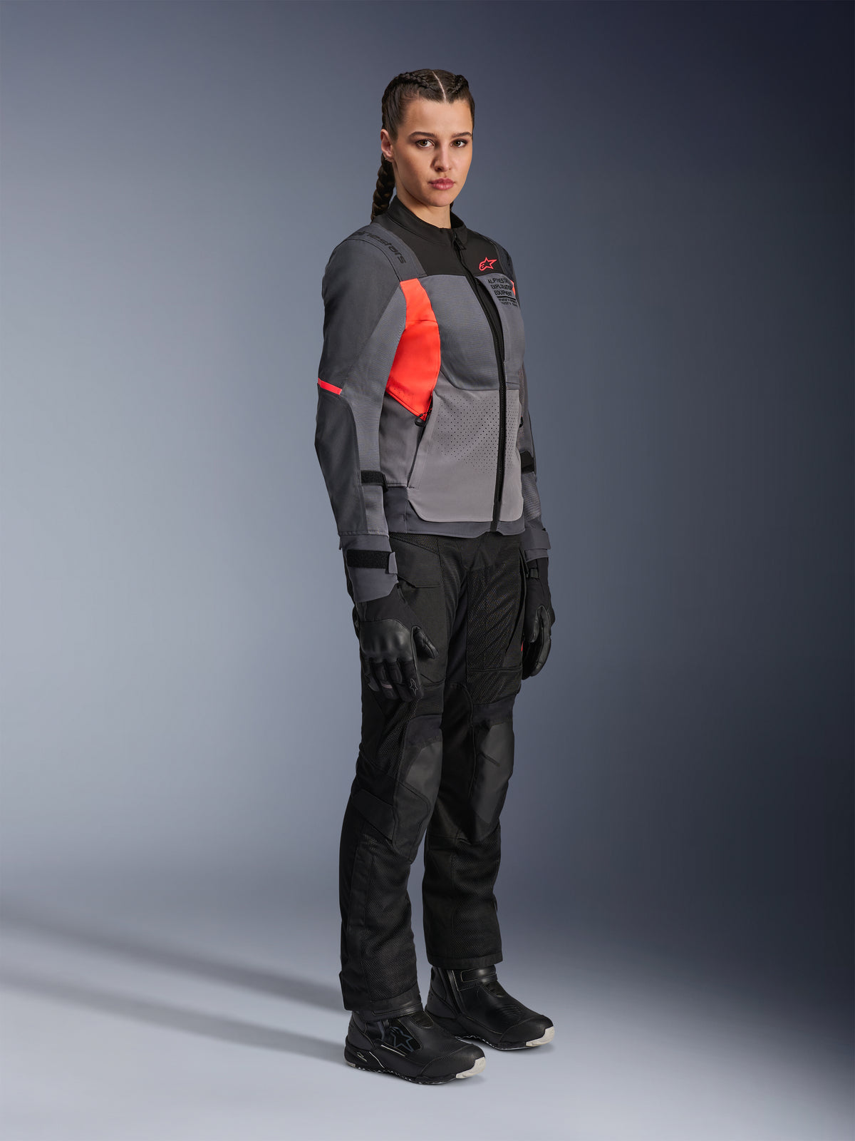 Motorradfahrerin in der Alpinestars Stella ST-2 Air Jacke in Deep Gray, Dark Gray und Fluo Coral, kombiniert mit einer schwarzen Motorradhose, Handschuhen und Stiefeln, mit einem modernen Touring-Design, Mesh-Einsätzen und reflektierenden Details.