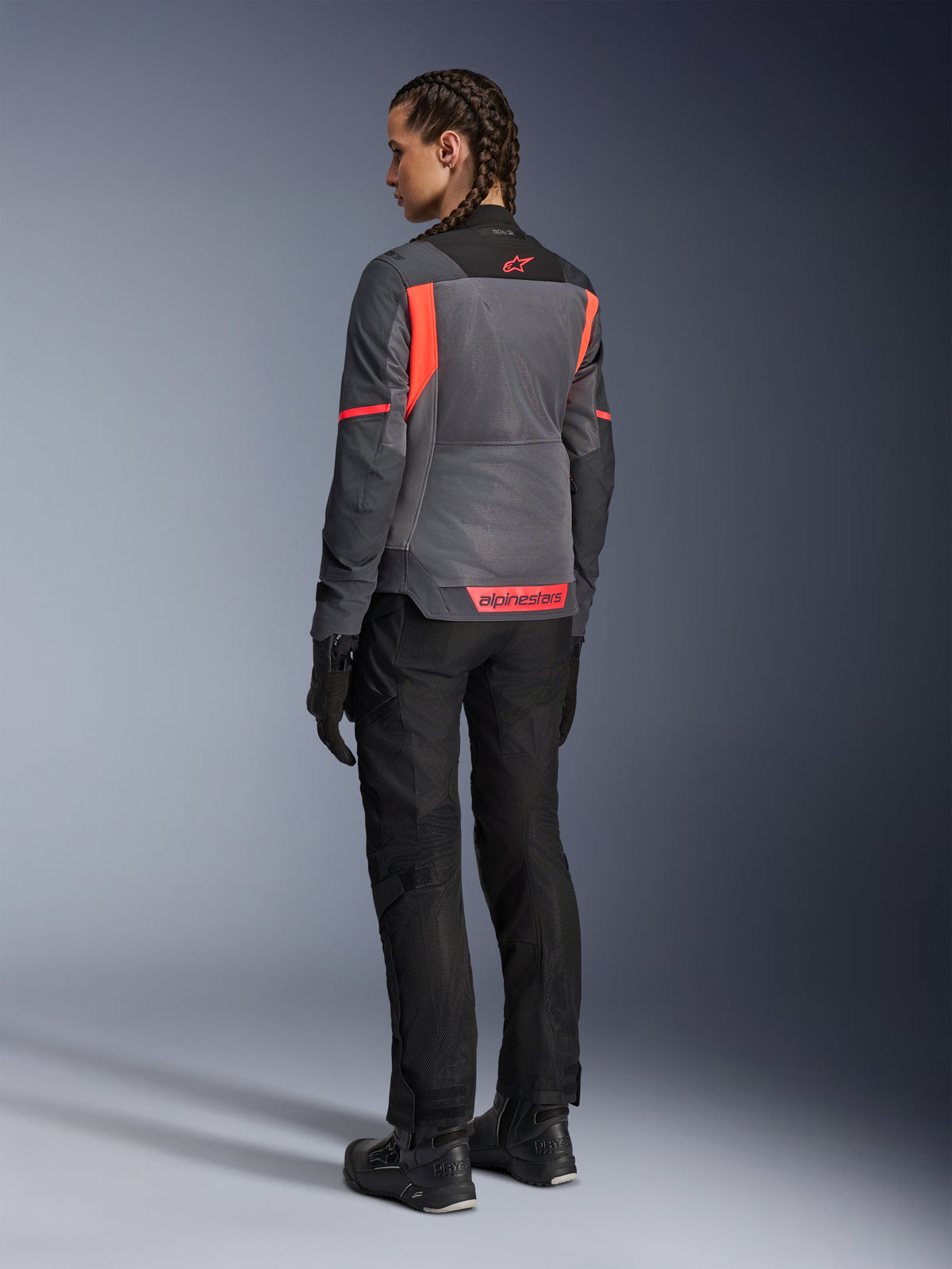 Frau in der Alpinestars Stella St-2 Air Jacke, Motorrad-Tourenjacke, Tiefgrau und Dunkelgrau mit Akzenten in Fluo Coral, kombiniert mit schwarzer Motorradhose und Handschuhen, Rückansicht mit großen Mesh-Einsätzen und reflektierendem Coral-Branding, konzipiert für sommerliche Tourenabenteuer
