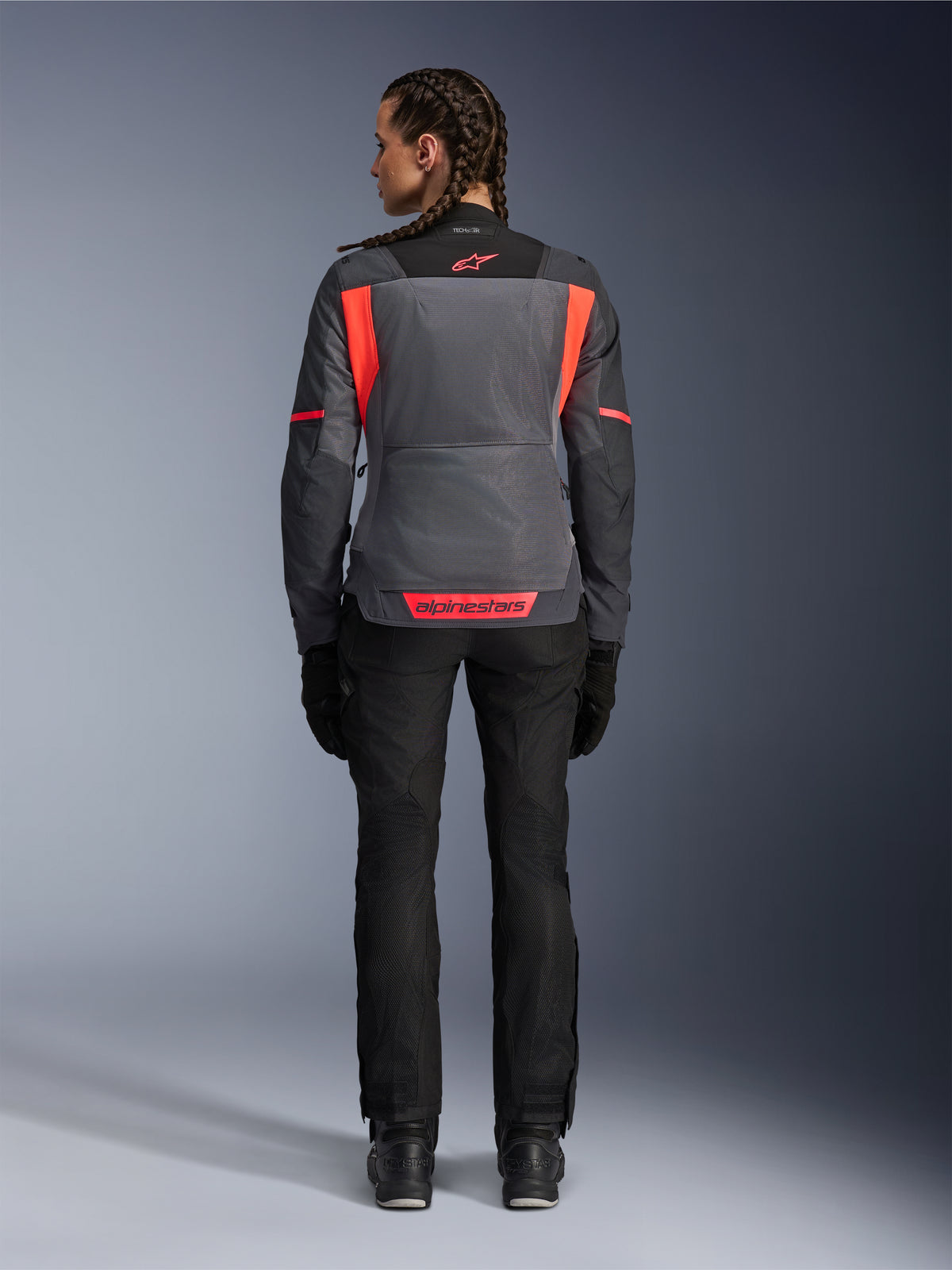 Rückansicht eines Models in der Alpinestars Stella ST-2 Air Damenjacke in Deep Gray und Dark Gray mit Fluo Coral Akzenten, mit großen Mesh-Belüftungseinsätzen am Rücken und reflektierenden Logo-Details, kombiniert mit einer schwarzen Motorradhose und Handschuhen.