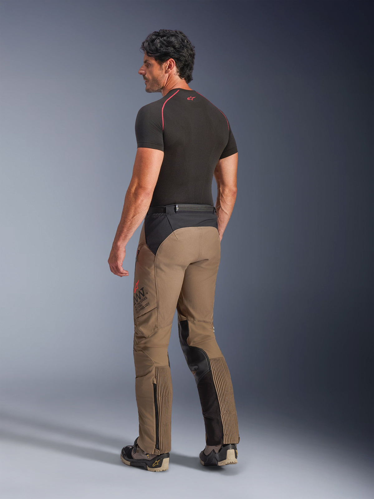 Alpinestars AMT-8 Stretch Drystar® XF Pants in Walnut Black, adventure-touring motorcycle pants shown from a back three-quarter view, featuring walnut tan textile with black panels on the seat and inner legs, accordion stretch calf panels, and AMT branding on the side, worn with a black base layer.