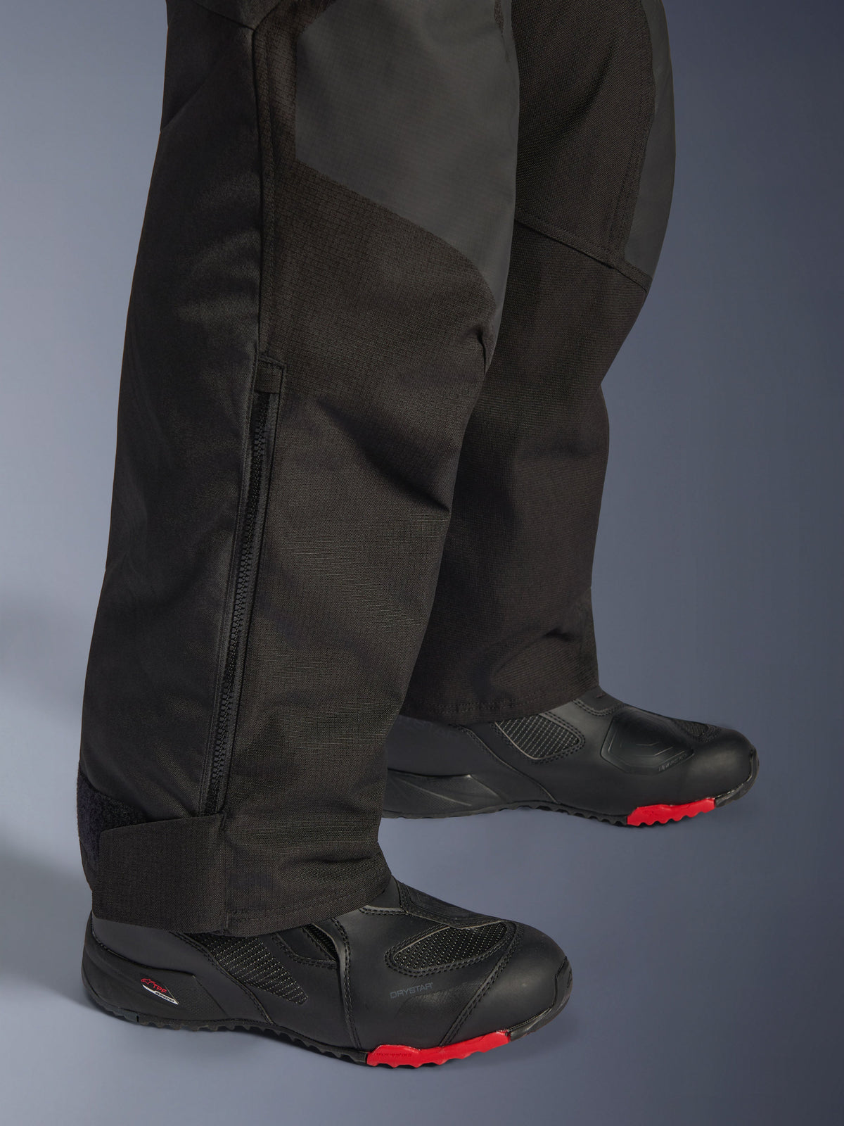 Alpinestars Andes V4 Drystar® Hose – Lang, Schwarze Motorrad-Abenteuerhose, Nahaufnahme des Unterschenkels mit verstellbarem Reißverschluss am Beinabschluss und strapazierfähiger Textilkonstruktion, getragen mit schwarzen Motorradstiefeln mit roten Akzenten