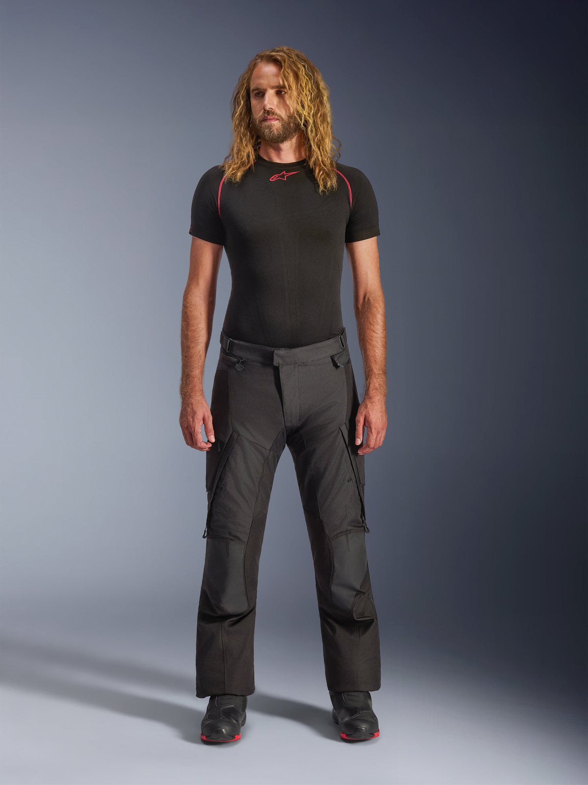 Männliches Model trägt Alpinestars Andes V4 Drystar® Hose - Lang in Schwarz, Adventure-Touring-Motorradhose mit verstärkten Knieeinsätzen und Cargotaschen, kombiniert mit einem schwarzen kurzärmeligen Base Layer und schwarzen Motorradstiefeln.