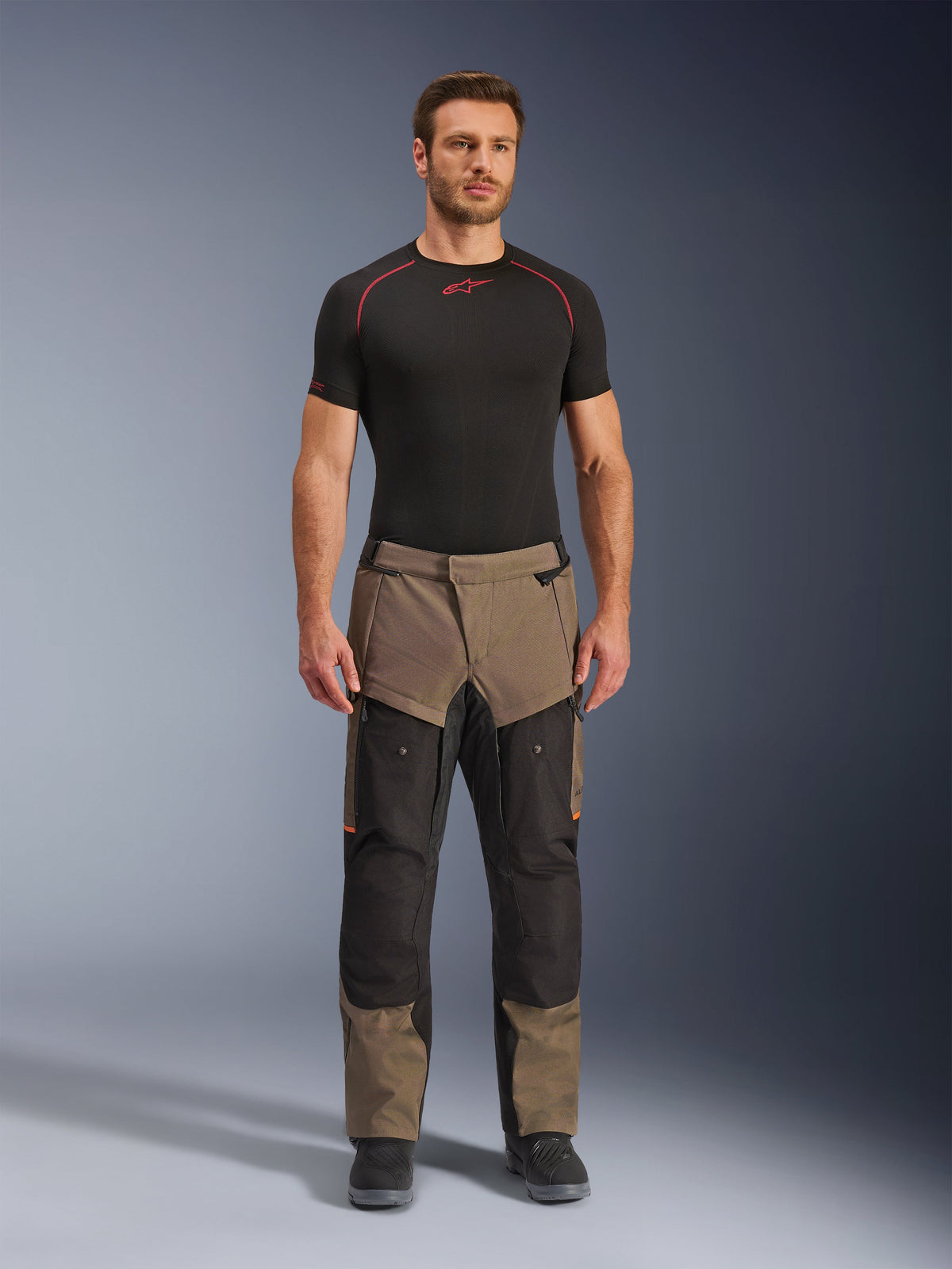 Alpinestars Cusco Drystar® Hose, Walnut Black, Adventure-Touring-Motorradhose, getragen von einem männlichen Model mit einem schwarzen Base-Layer und schwarzen Stiefeln, mit walnussfarbenen und schwarzen Einsätzen, orangefarbenen Akzenten und direkten Belüftungsreißverschlüssen an den Oberschenkeln.