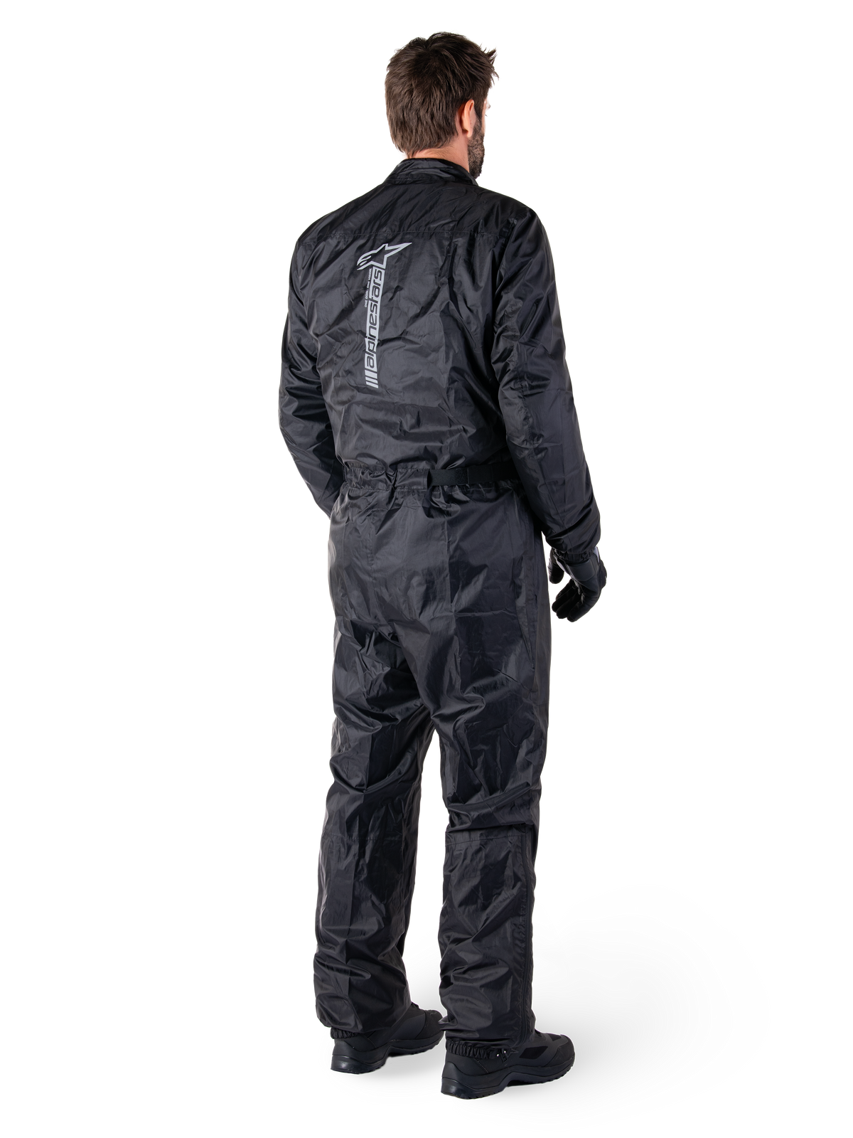 Alpinestars Hurricane Rain V2 Suit, schwarzer einteiliger wasserdichter Motorrad-Regenanzug, Rückansicht mit großem vertikalem reflektierendem Logo, leichtes windabweisendes Gewebe, getragen mit schwarzen Handschuhen und Stiefeln