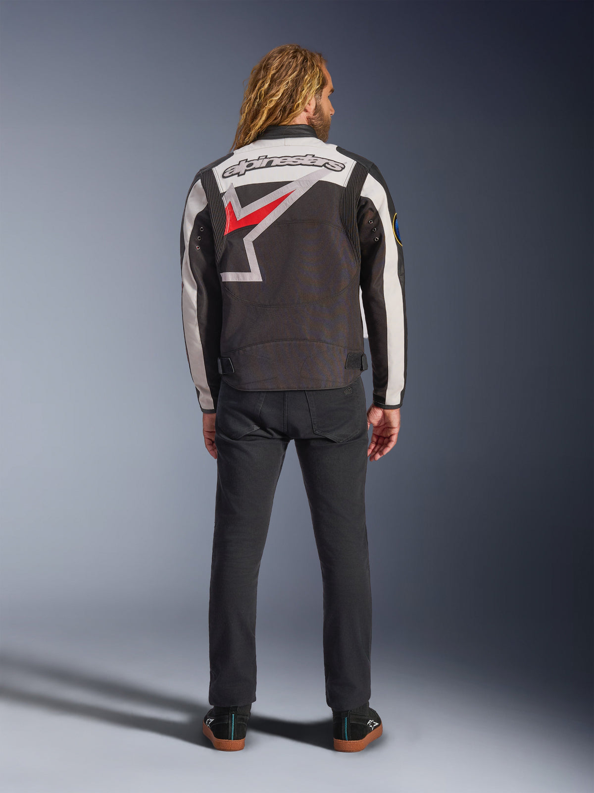 Alpinestars Axton Jacke, Motorradjacke, Schwarz, Off-White und Bright Red, Rückansicht an einem Model, mit einem großen Alpinestars-Sternlogo auf dem Rücken, Off-White-Ärmeleinsätzen, getragen mit einer schwarzen Hose, von Vintage-Rennsportbekleidung inspiriertes Design