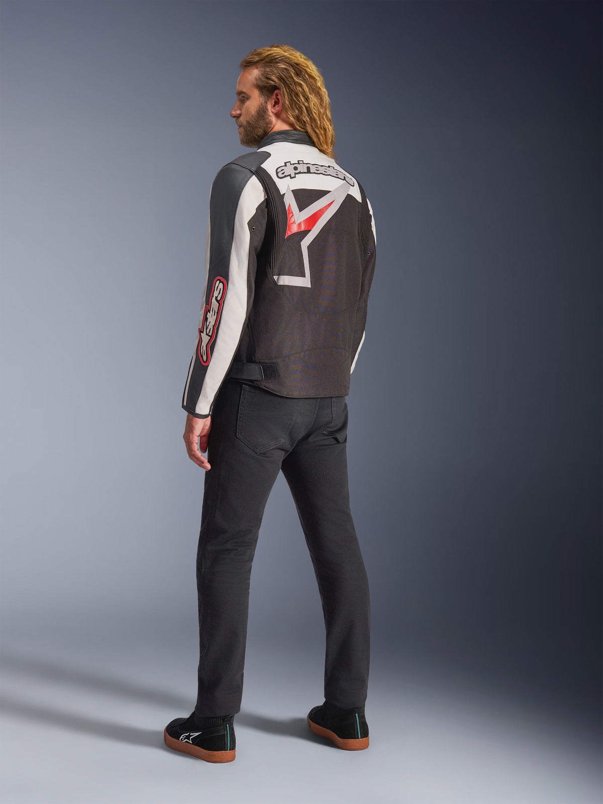 Alpinestars Axton Jacke, Motorradjacke, Schwarz, Off-White und leuchtendes Rot, Rückansicht getragen von einem männlichen Model, mit großem Alpinestars Stern-Logo und Schriftzug auf dem Rücken, Vintage-Rennsport-inspiriertes Design mit Akkordeon-Einsätzen, kombiniert mit schwarzer Hose und Sneakern