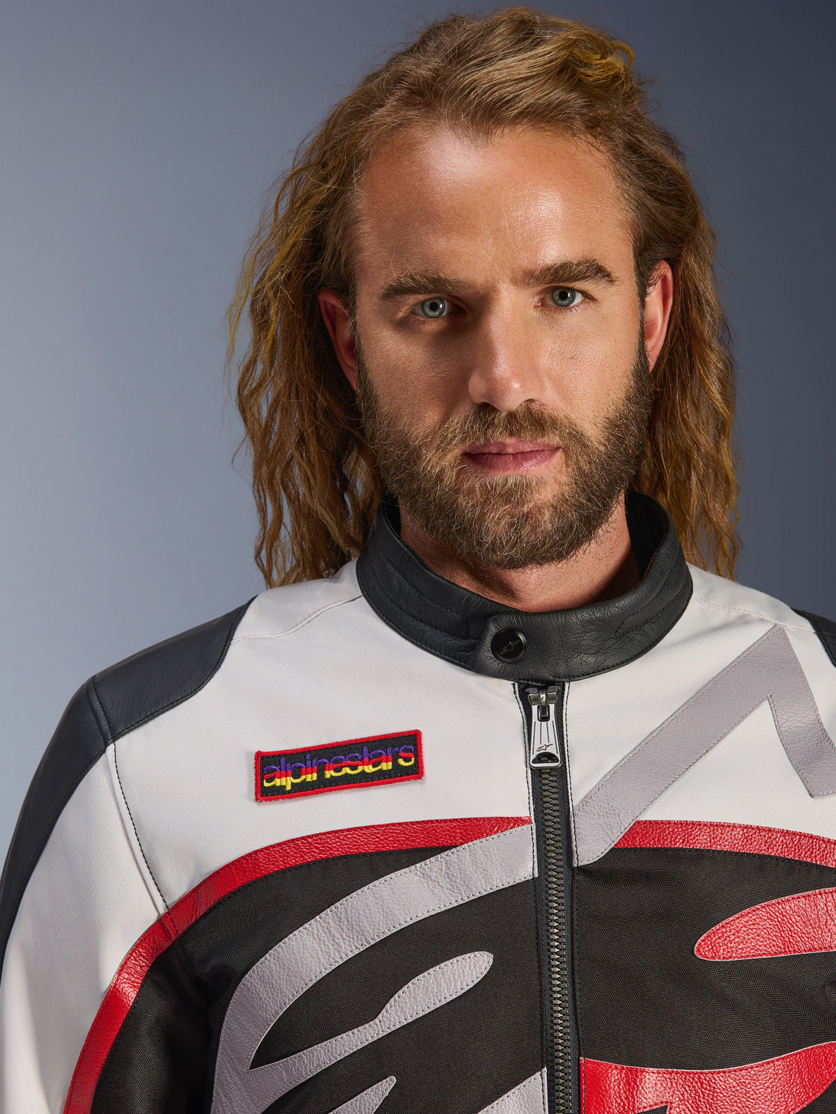 Mann in der Alpinestars Axton Jacke, Vintage-inspirierte Motorradjacke in Schwarz, Off-White und Bright Red, Nahaufnahme mit Marken-Brustaufnäher und minimalistischem Biker-Kragen, zeitgemäße Straßenfahrausrüstung
