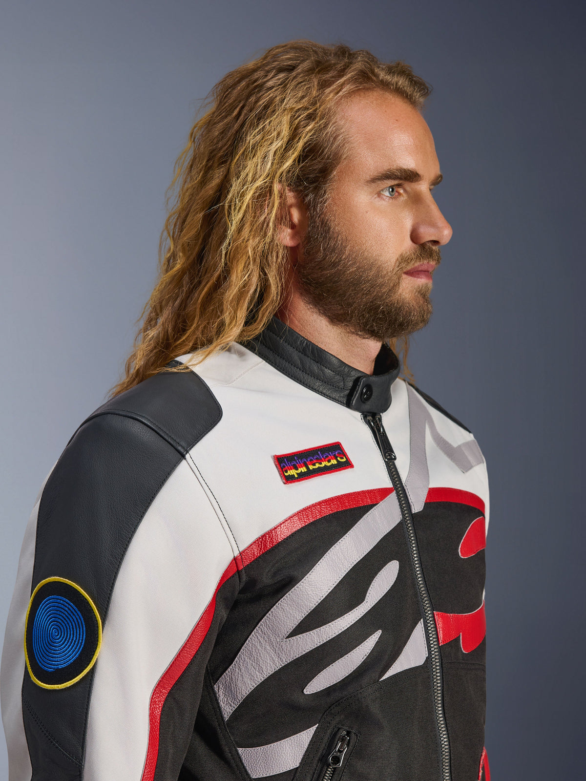 Mann mit langen Haaren und Bart trägt die Alpinestars Axton Jacke, Motorradjacke, Schwarz, Off-White und Bright Red, vom Vintage-Rennsport inspiriertes Design mit Marken-Patches und umlaufendem Logo, Nahaufnahme in der Profilansicht, urbaner zeitgenössischer Stil