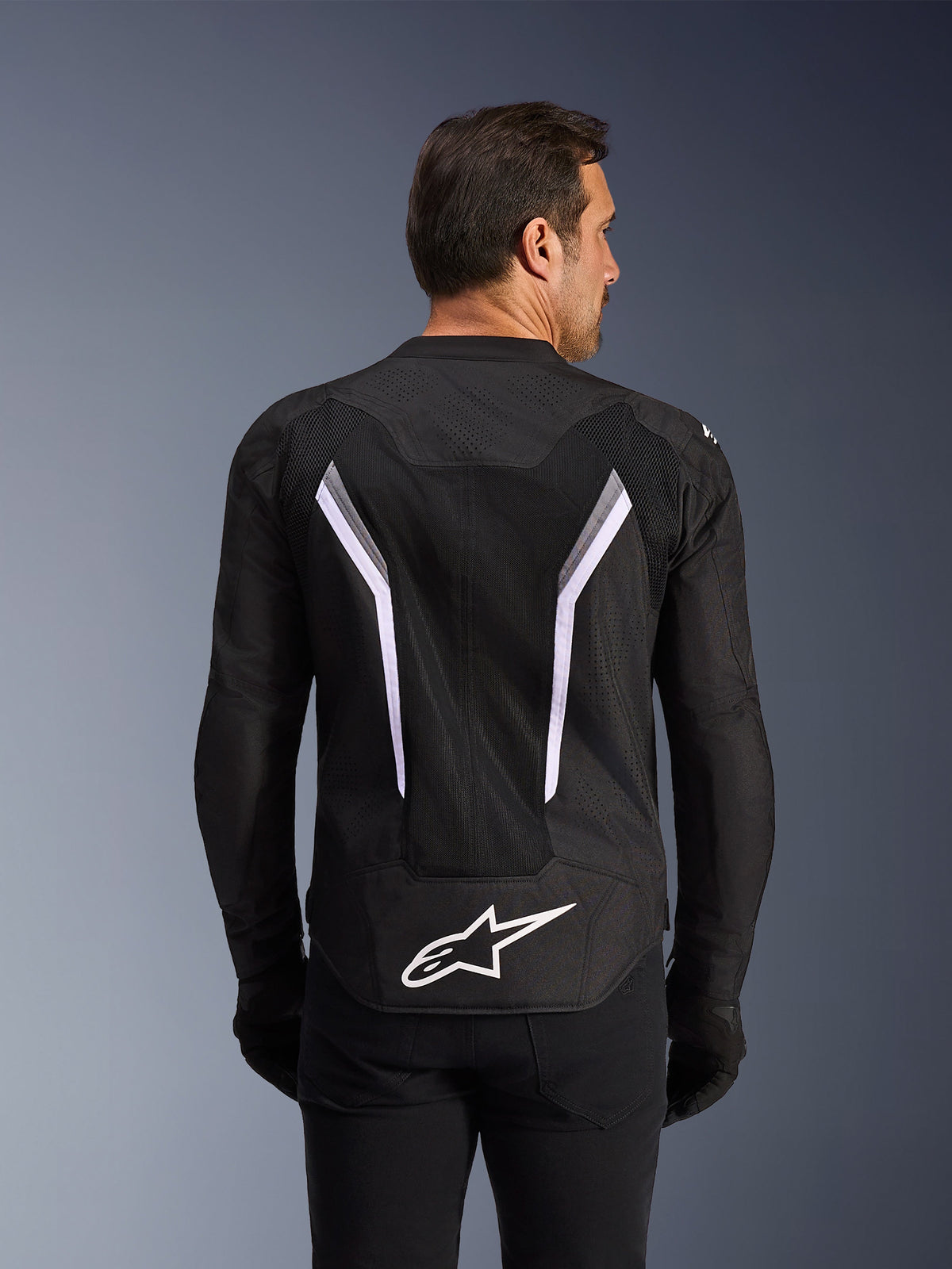 Alpinestars AST V3 Air Jacke, Motorradjacke, Schwarz mit weißen und dunkelgrauen Akzenten, Rückansicht mit umfangreichen Mesh-Belüftungseinsätzen, großen weißen V-förmigen Streifen und weißem Alpinestars-Logo am unteren Rücken, getragen mit schwarzen Handschuhen und Hosen