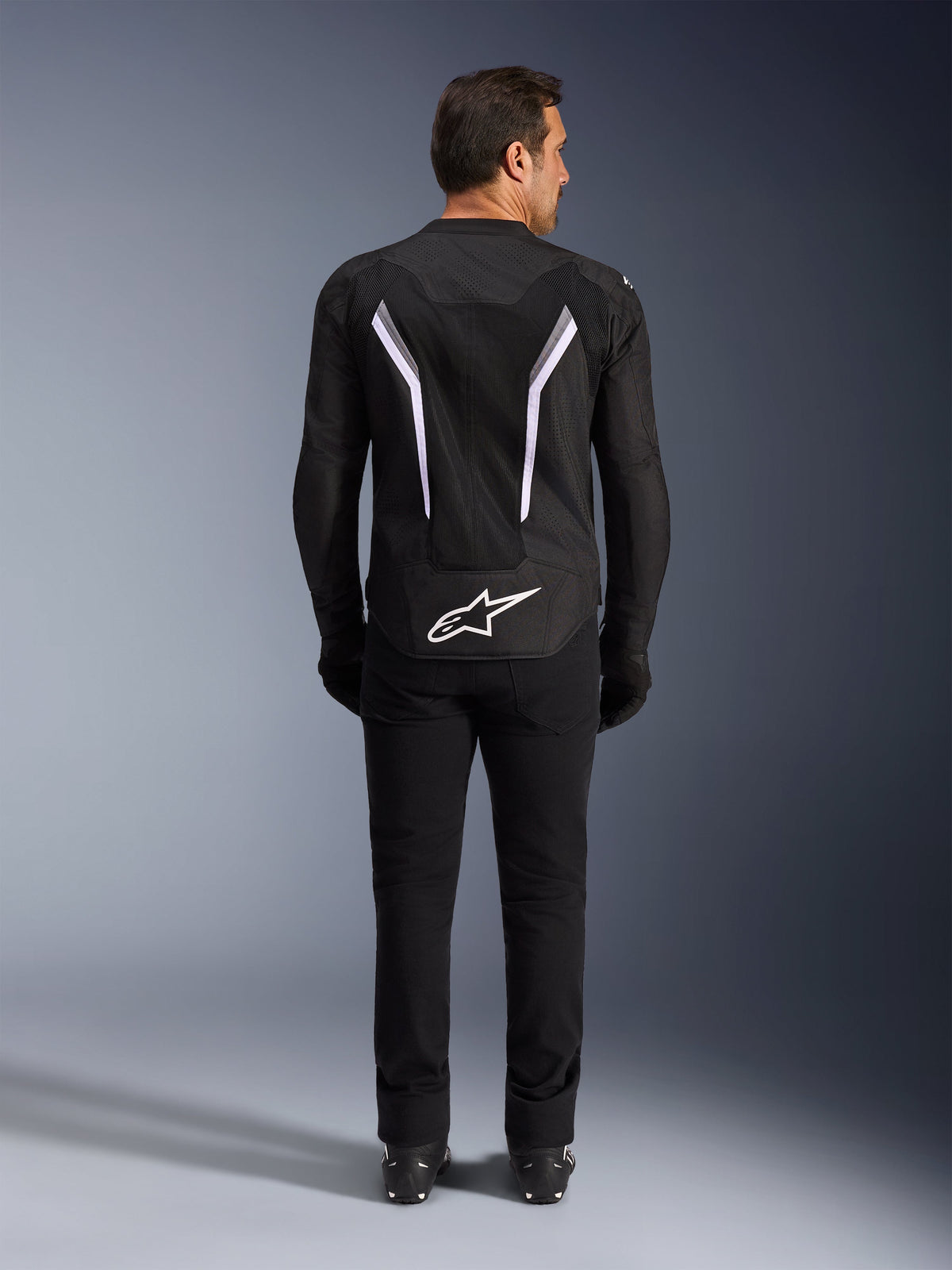 Alpinestars AST V3 Air Jacke, Rückansicht, Schwarz und Dunkelgrau mit weißen Akzenten, großes weißes Astar-Logo am unteren Rücken, reflektierende Streifen, Mesh-Textil-Konstruktion, sportlicher Look mit schwarzer Hose und Stiefeln