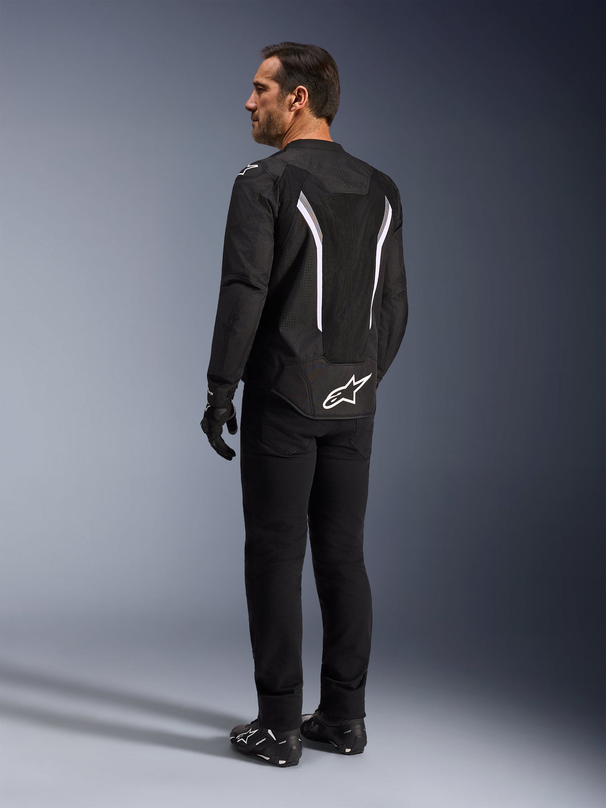 Alpinestars AST V3 Air Jacke, schwarz-weiße Motorradjacke, Rückansicht am Modell, mit Mesheinsätzen für Luftzirkulation und weißem Logo am unteren Rücken, kombiniert mit schwarzer technischer Fahrerausrüstung