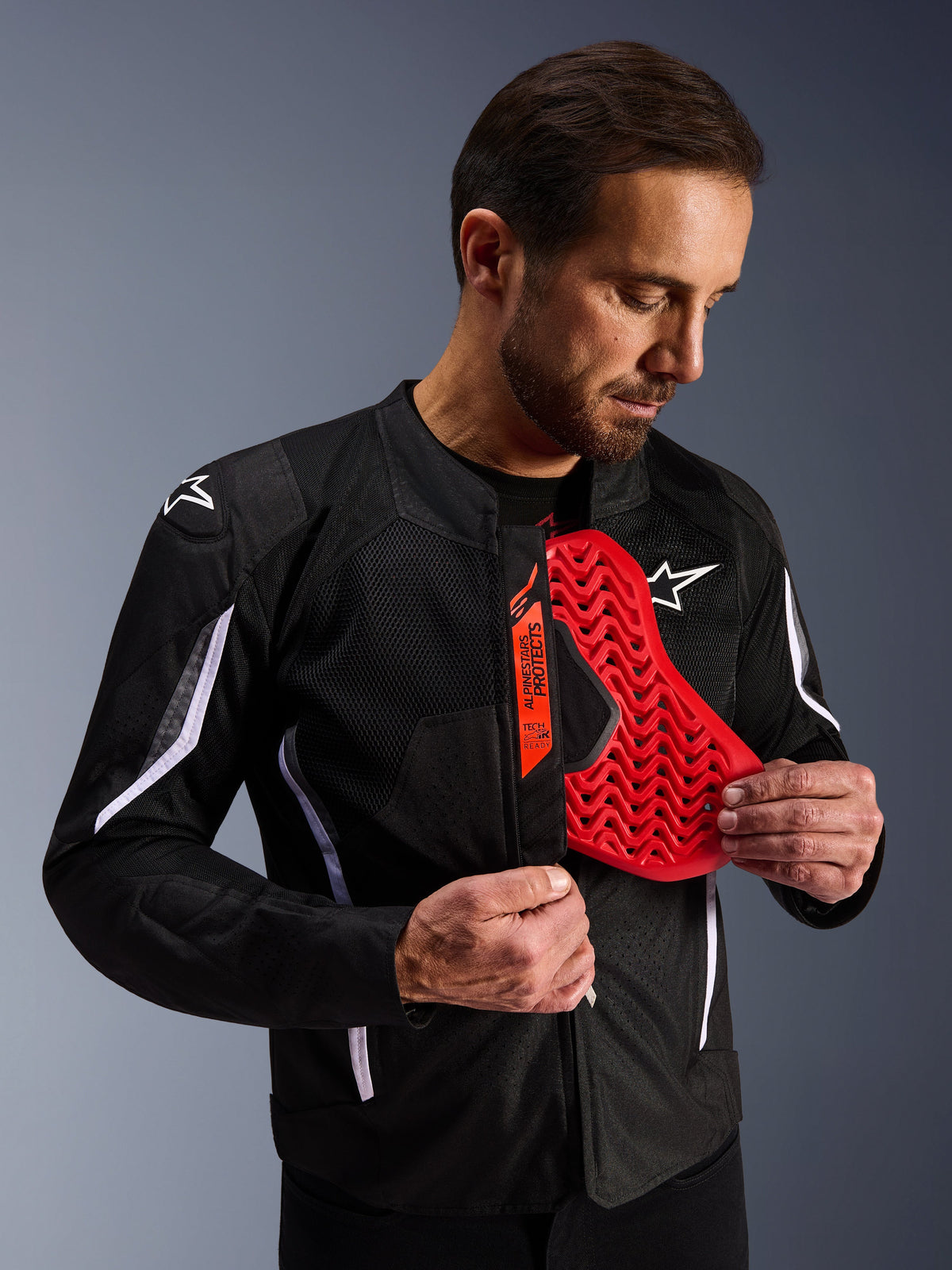 Alpinestars AST V3 Air Jacke in Schwarz, Mann zeigt einen leuchtend roten Brustprotektor-Einsatz mit Wabenmuster, Hervorhebung der Tech-Air Ready Kompatibilität und des integrierten internen Schutzes für Motorradsicherheit.