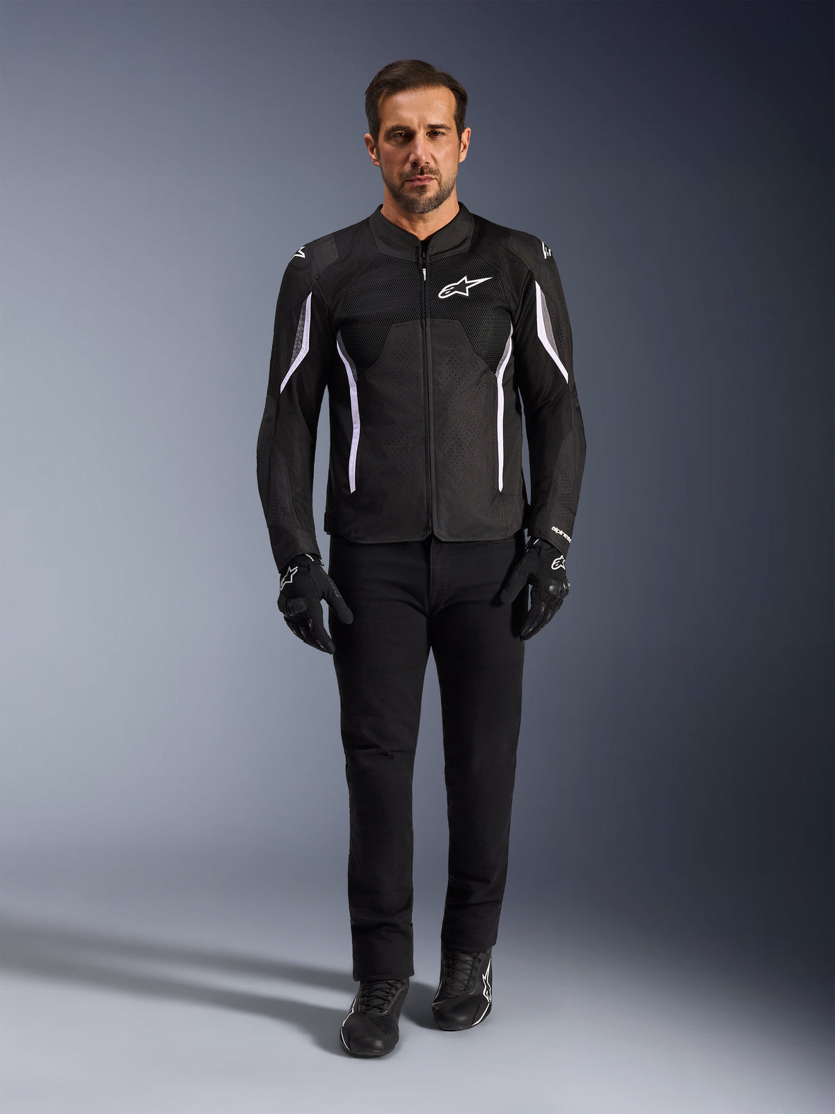 Männliches Model trägt die Alpinestars AST V3 Air Jacke in Schwarz mit weißen Akzenten, Mesh-Belüftungseinsätze und sportliche Passform, kombiniert mit schwarzer technischer Motorradhose, Handschuhen und Stiefeln, Vorderansicht, technische Motorradausrüstung für Fahrten bei warmem Wetter.