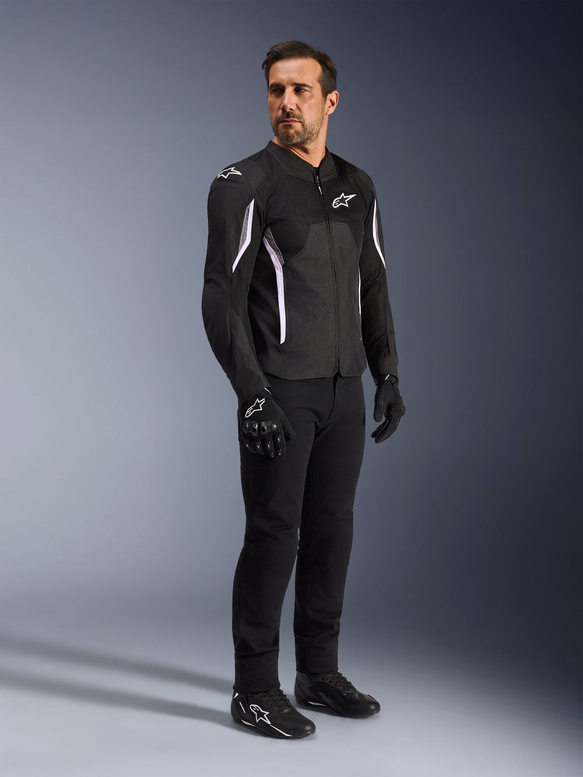 Alpinestars AST V3 Air Jacke, Schwarz mit weißen Akzenten und Branding, leichte Motorradjacke mit Mesh-Belüftung, getragen von einem Mann mit schwarzer Motorradhose, Handschuhen und Schuhen, technische Sommer-Fahrausrüstung für Sport und Straße.