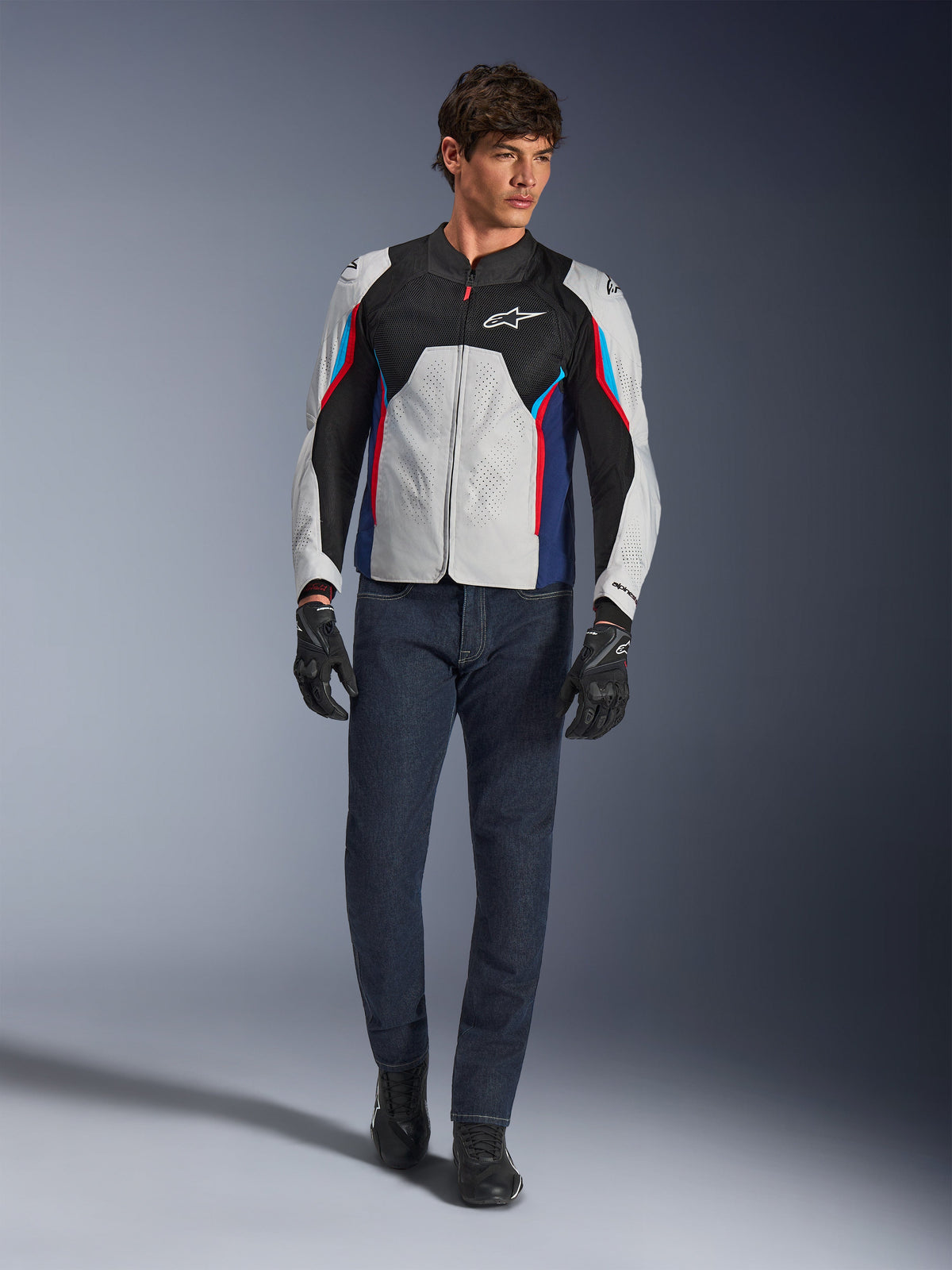 Male model wearing the Alpinestars AST V3 Air Jacket in Silver Deep Blue Bright Red, featuring mesh ventilation and perforated panels, paired with dark denim riding jeans and black gloves, sport-optimized motorcycle gear for warm weather.