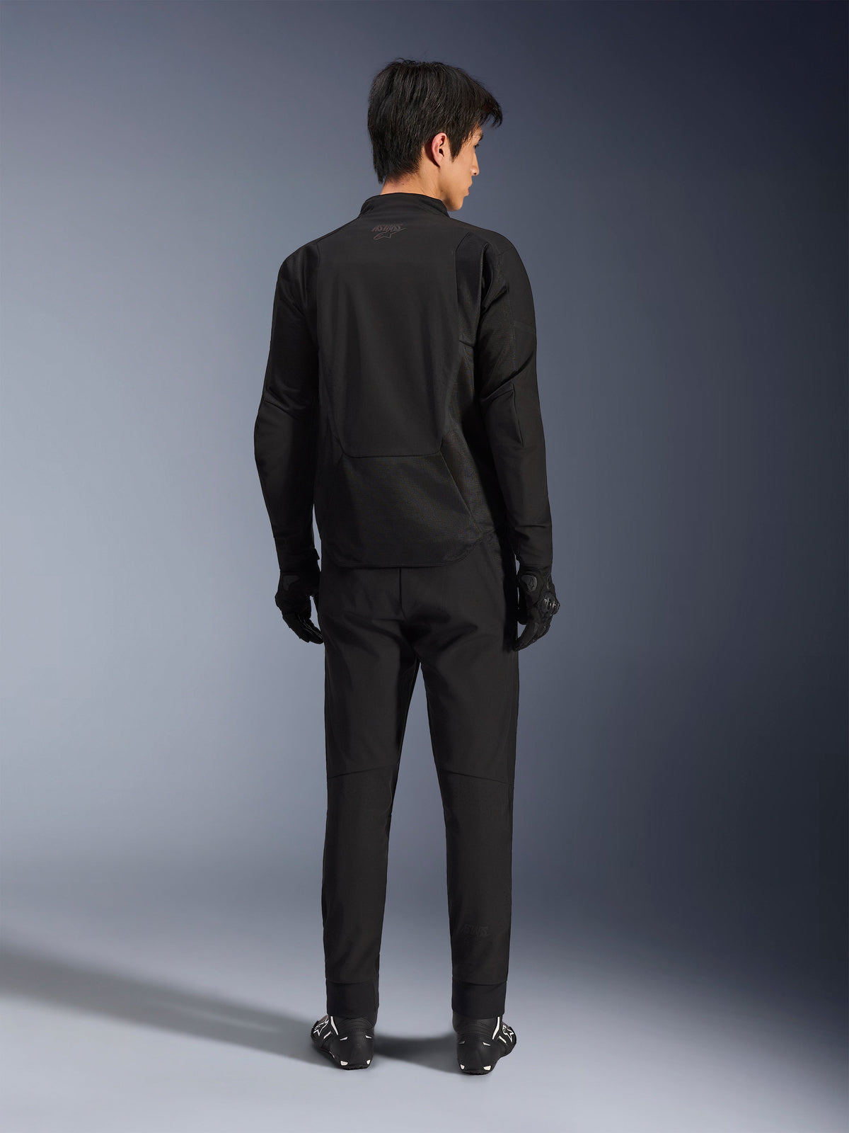 Alpinestars Aeroshell Airflow Jacke in Black Anthracite, Rückansicht eines männlichen Models in kompletter Motorradbekleidung inklusive schwarzer Hose und Handschuhen, mit einer leichten Softshell-Konstruktion und einem verlängerten Rücken für optimale Abdeckung.
