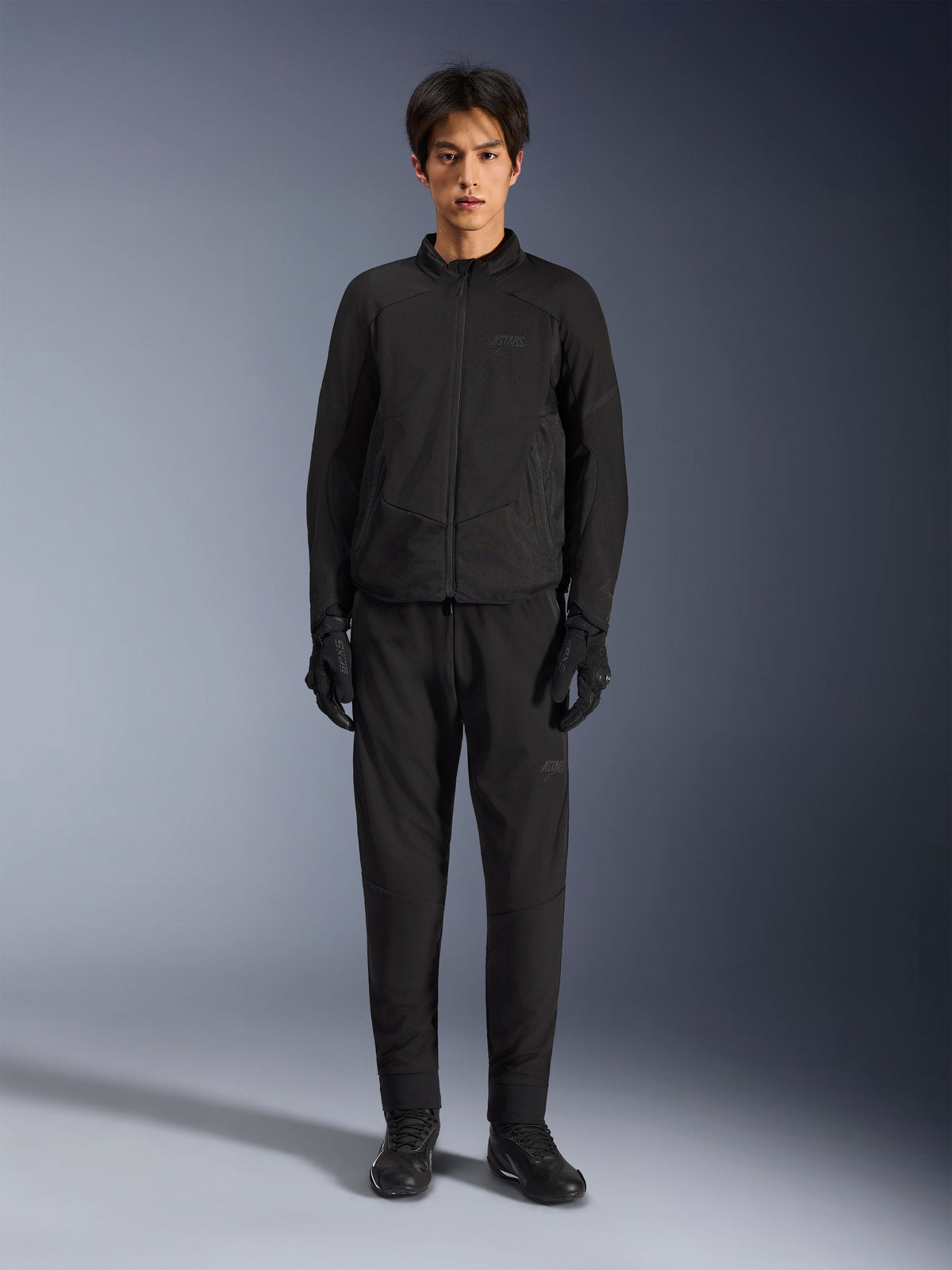 Alpinestars Aeroshell Airflow Jacke, Schwarze Motorradjacke, von Model getragen mit passender schwarzer Hose und Handschuhen, laserperforierte Brust und seitliche Mesh-Einsätze, sportliche Passform für Performance bei warmem Wetter