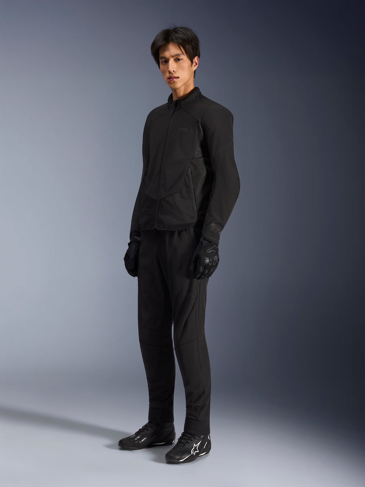 Alpinestars Aeroshell Airflow Jacke, Motorradjacke in Black Anthracite, mit laserperforierter Belüftung und Mesh-Einsätzen für den Luftstrom, getragen von einem männlichen Model mit passender schwarzer Fahrhose, Handschuhen und schwarzen Stiefeln mit weißen Logos, sportlich geschnittenes Design für Performance auf der Straße