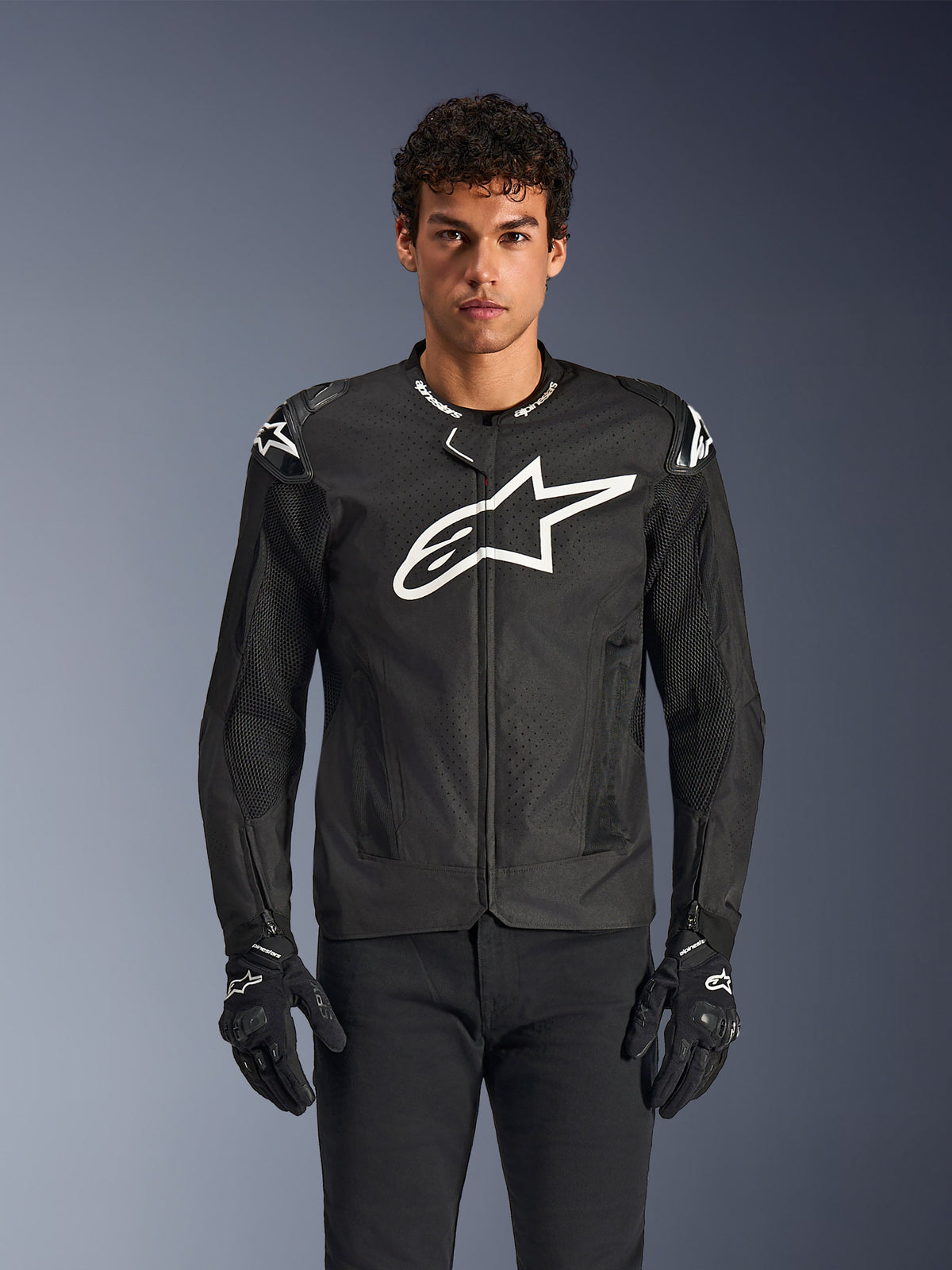 Alpinestars T-GP Air Jacket, Black textile motorcycle jacket with a large white star logo on the chest, features perforated ventilation panels and 3D mesh, external shoulder sliders, worn with black and white gloves, sleek sporty design for warm-weather road riding