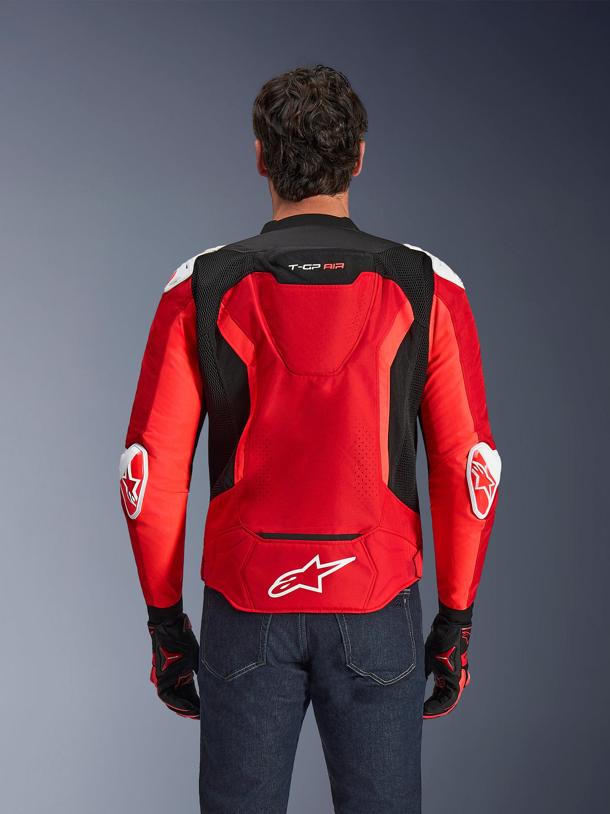 Alpinestars T-GP Air Jacket, Textile Motorcycle Jacket, Bright Red and Black with white accents, back view showing the aero hump and mesh ventilation panels, featuring the Alpinestars star logo and T-GP Air branding, worn with dark jeans and racing gloves.
